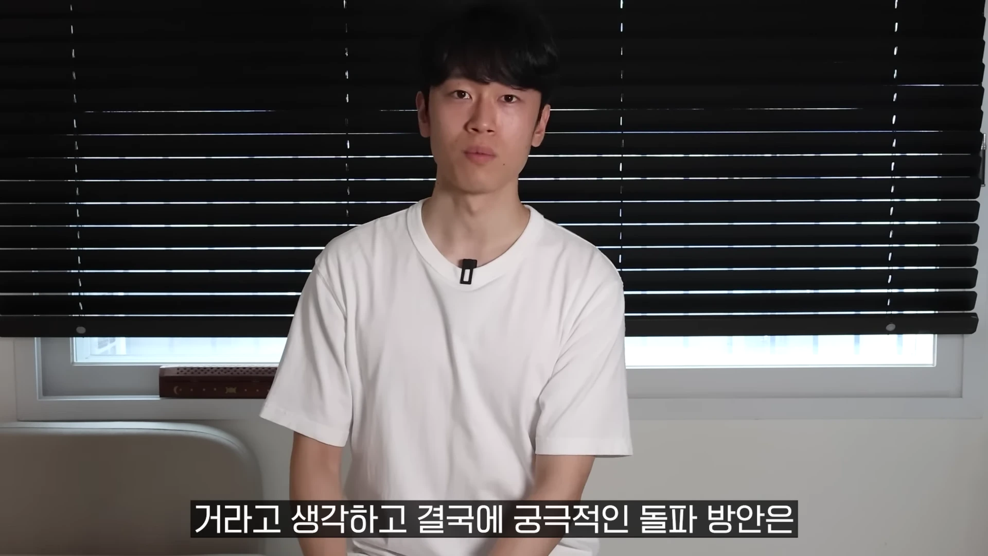 흰색 반소매 티셔츠를 입고 검은색 블라인드 창문 앞에 앉아 카메라를 응시하는 젊은 남성이 화면 중앙에 보입니다.