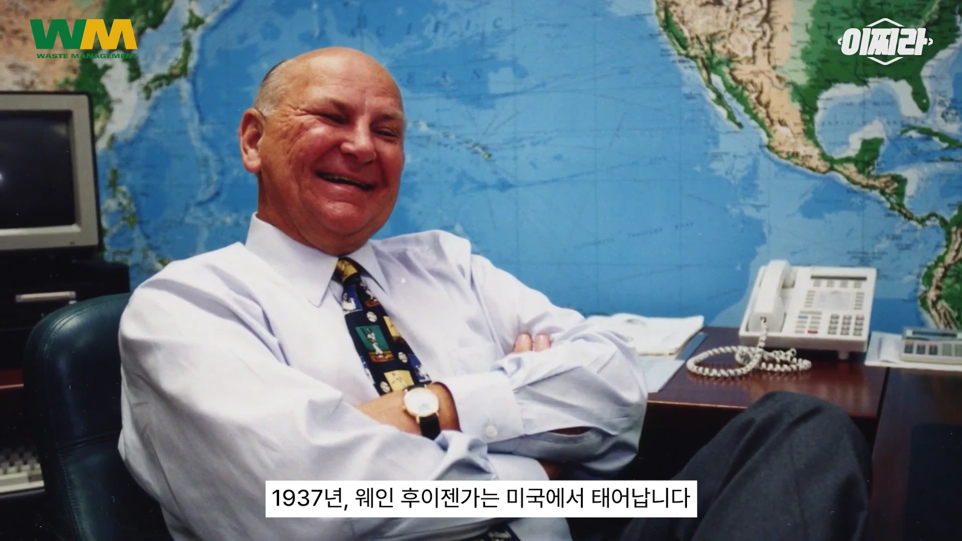 세계 지도가 그려진 벽을 배경으로 사무실 의자에 앉아 밝게 웃고 있는 중년 남성의 모습이며 아래에는 한국어 자막이 흐릅니다.