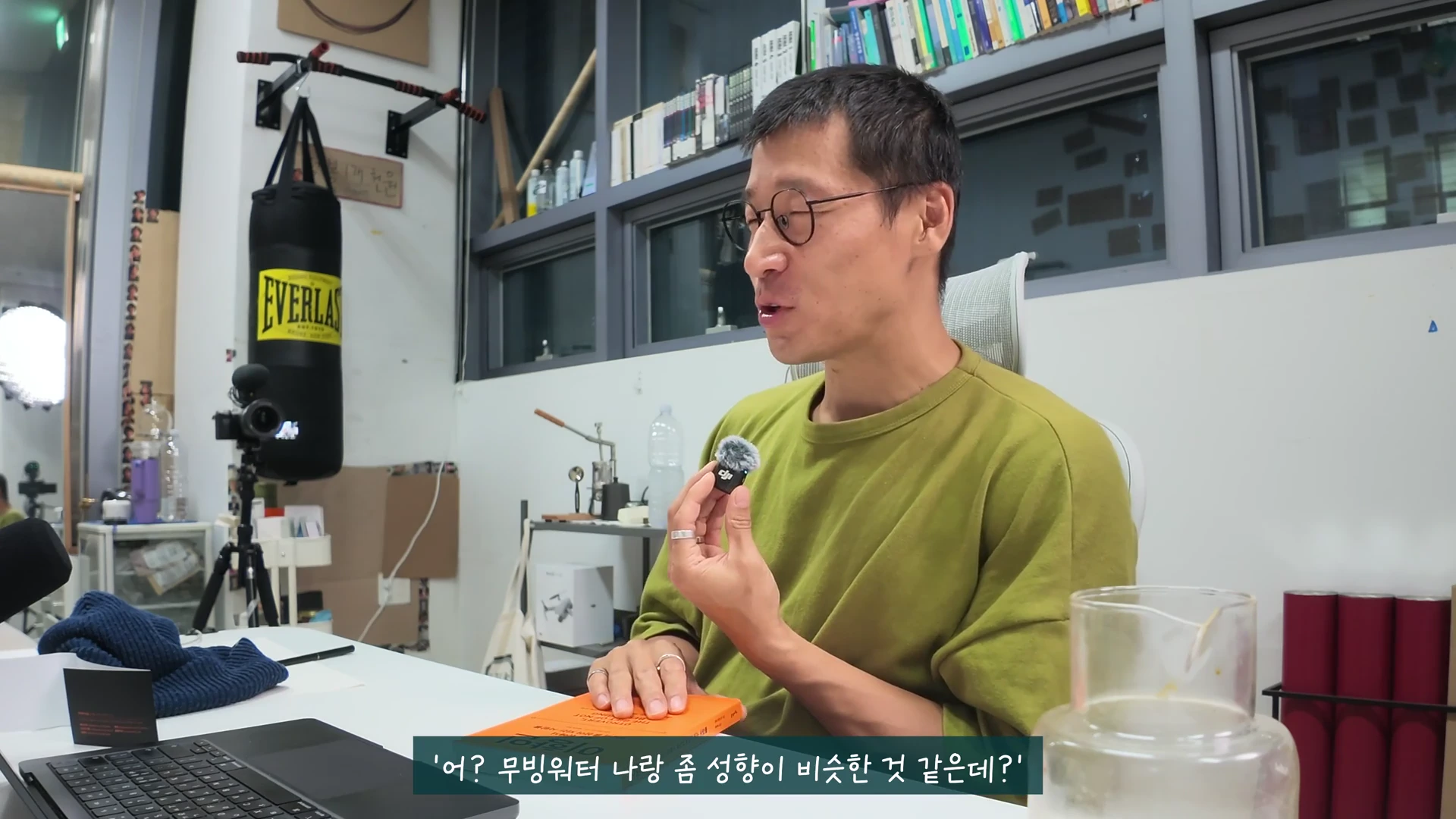 안경을 쓴 남성이 올리브색 티셔츠를 입고 책상에 앉아 손에 작은 마이크를 든 채 이야기하고 있는 모습