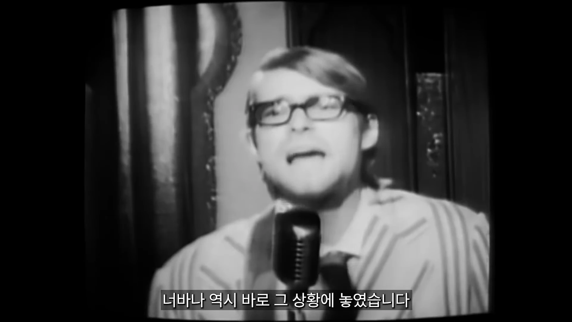 흑백 영상 속에서 안경을 쓴 남성이 마이크 앞에서 노래하거나 이야기하고 있으며, 화면 하단에는 한국어 자막이 표시되어 있습니다.
