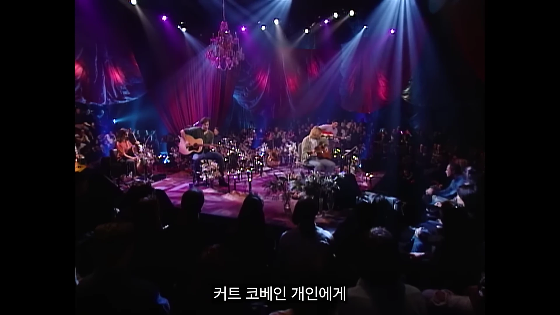 조명이 화려하게 비치는 실내 무대 위에서 통기타를 연주하며 노래하는 밴드 멤버들과 이를 지켜보는 관객들의 모습이 담긴 공연 장면.