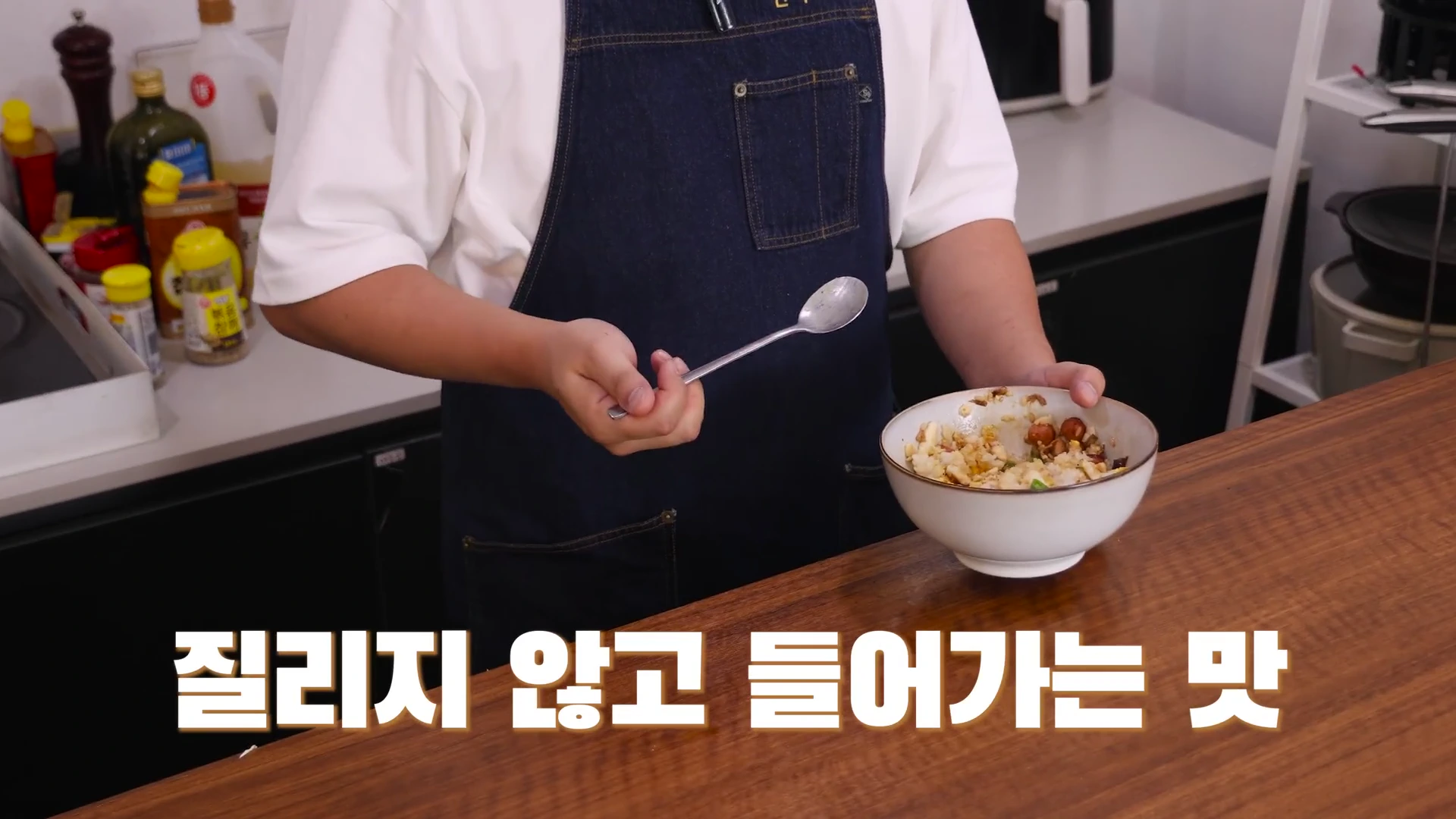 앞치마를 입은 요리사가 한 손에는 숟가락을, 다른 한 손에는 완성된 덮밥 그릇을 들고 있는 모습. 화면 하단에는 흰색 자막이 적혀 있음.