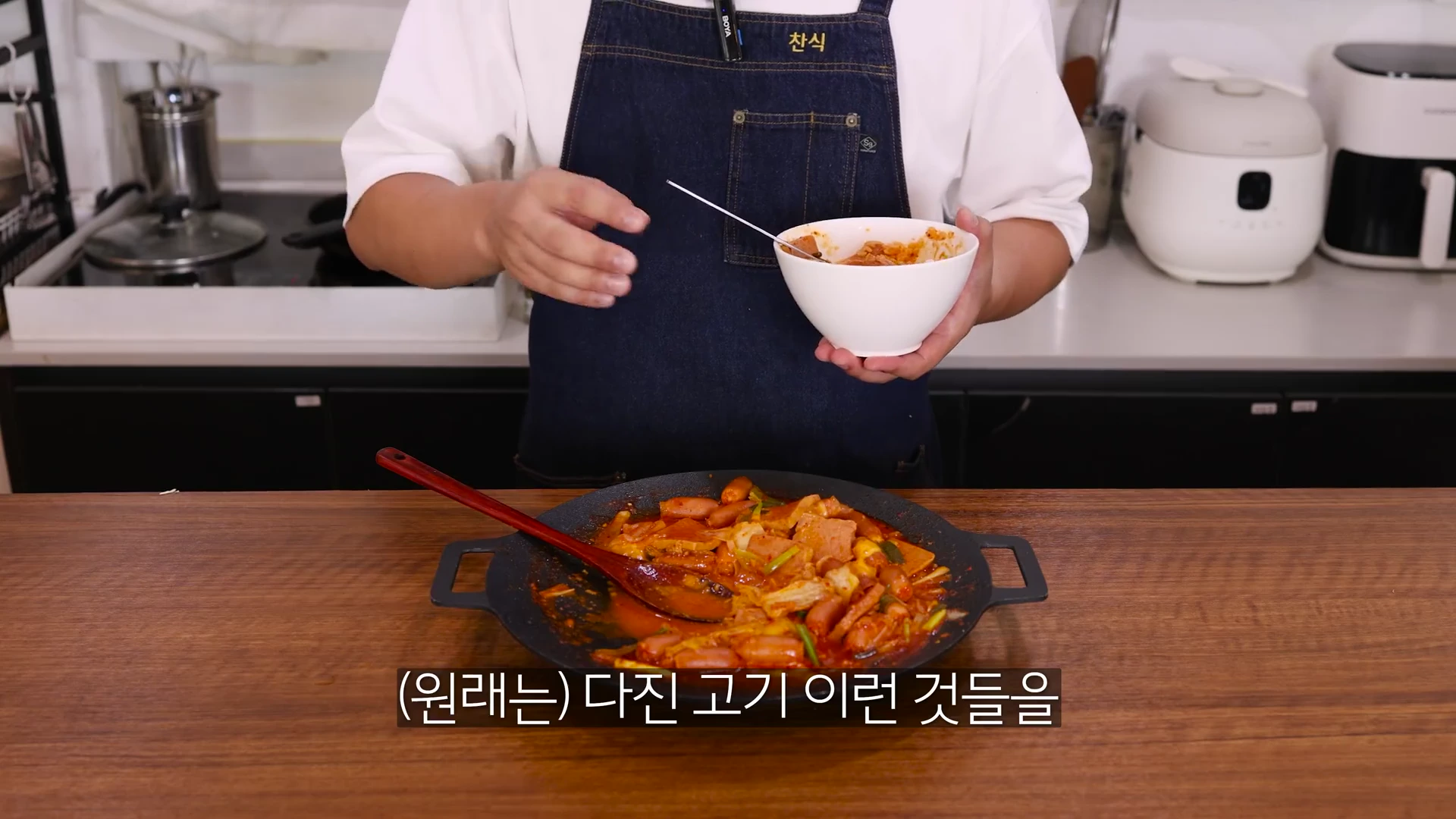 앞치마를 두른 요리사가 한 손에는 완성된 요리가 담긴 그릇을 들고 다른 한 손으로는 요리에 대해 설명하고 있으며, 앞쪽에는 비엔나소시지와 햄, 김치 등이 들어간 붉은 양념의 부대볶음이 담긴 프라이팬이 놓여 있다.