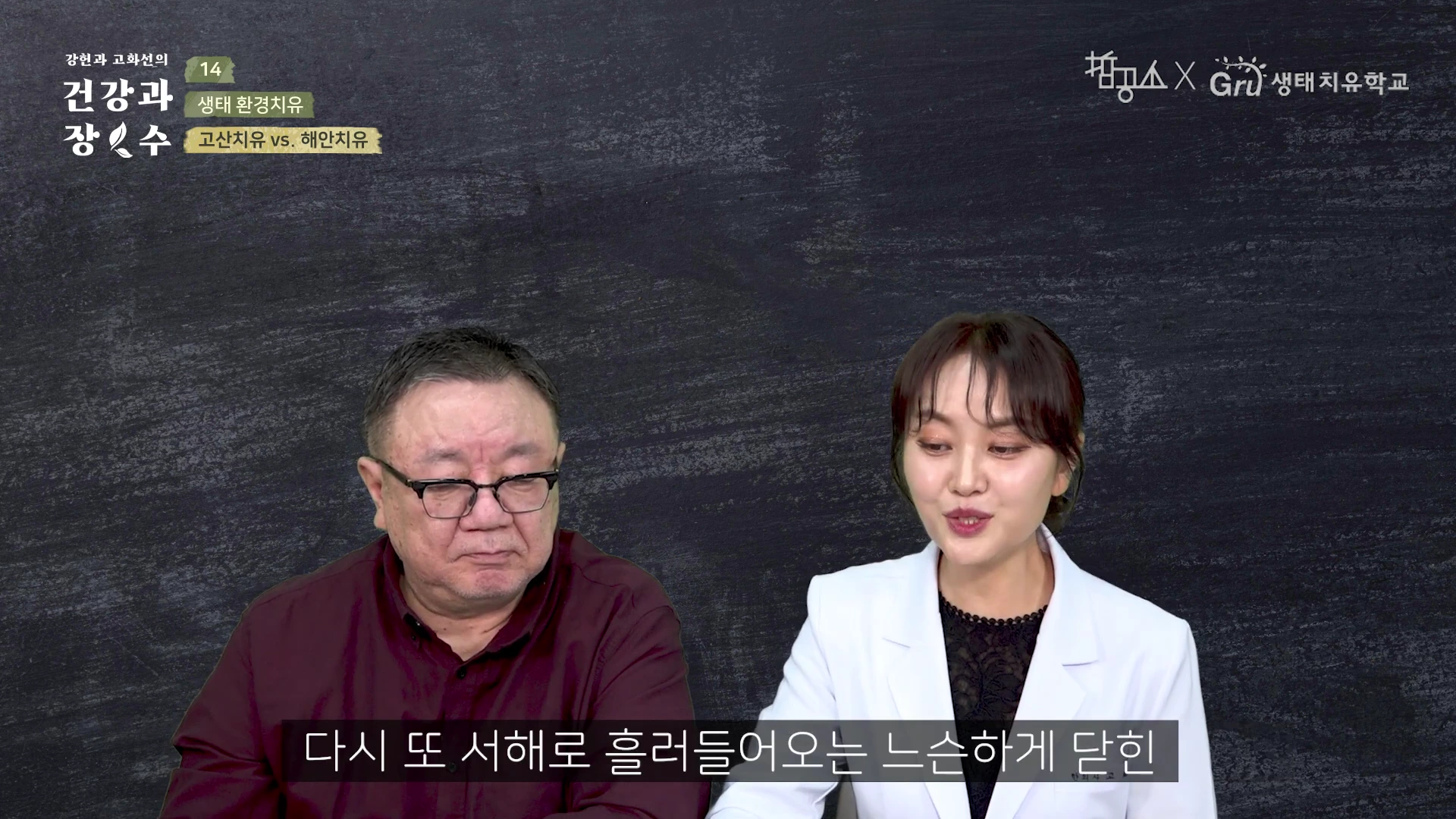 두 사람이 어두운 배경 앞에서 대화를 나누고 있는 스튜디오 장면으로, 왼쪽에는 안경을 쓴 남성이 있고 오른쪽에는 흰색 재킷을 입은 여성이 있습니다.