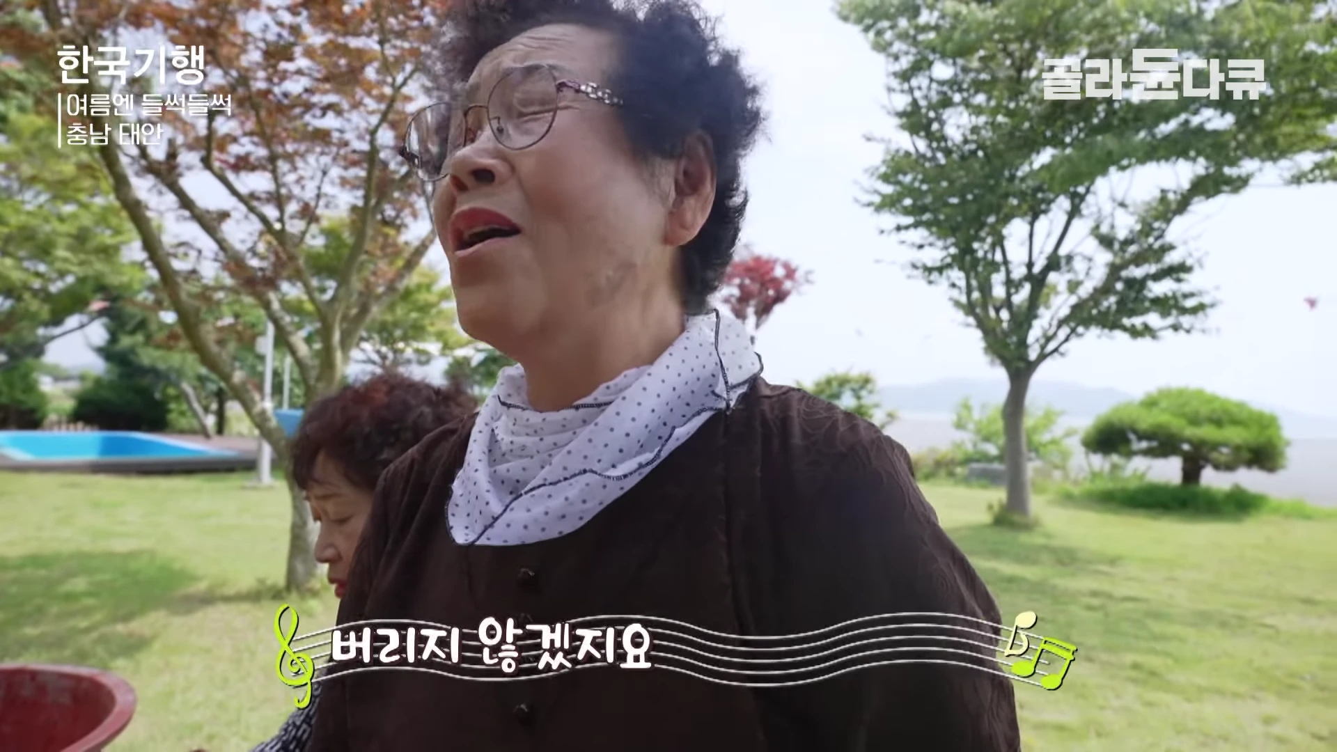 안경을 쓴 고령의 여성이 야외에서 노래를 부르고 있고, 뒤쪽으로 다른 여성이 서 있는 모습이 담긴 화면이다.