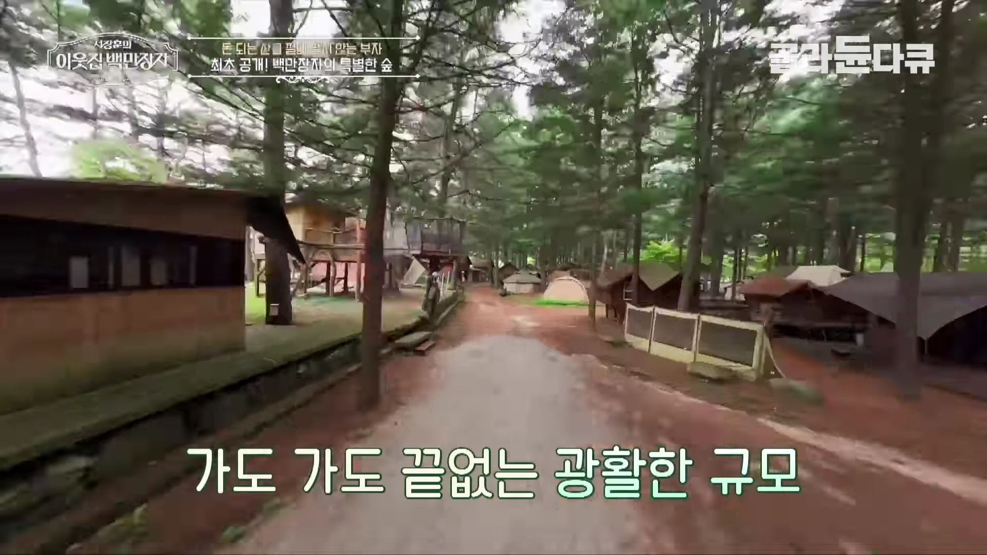숲속에 길게 뻗은 흙길과 양옆으로 세워진 여러 개의 오두막과 텐트들이 보이는 풍경