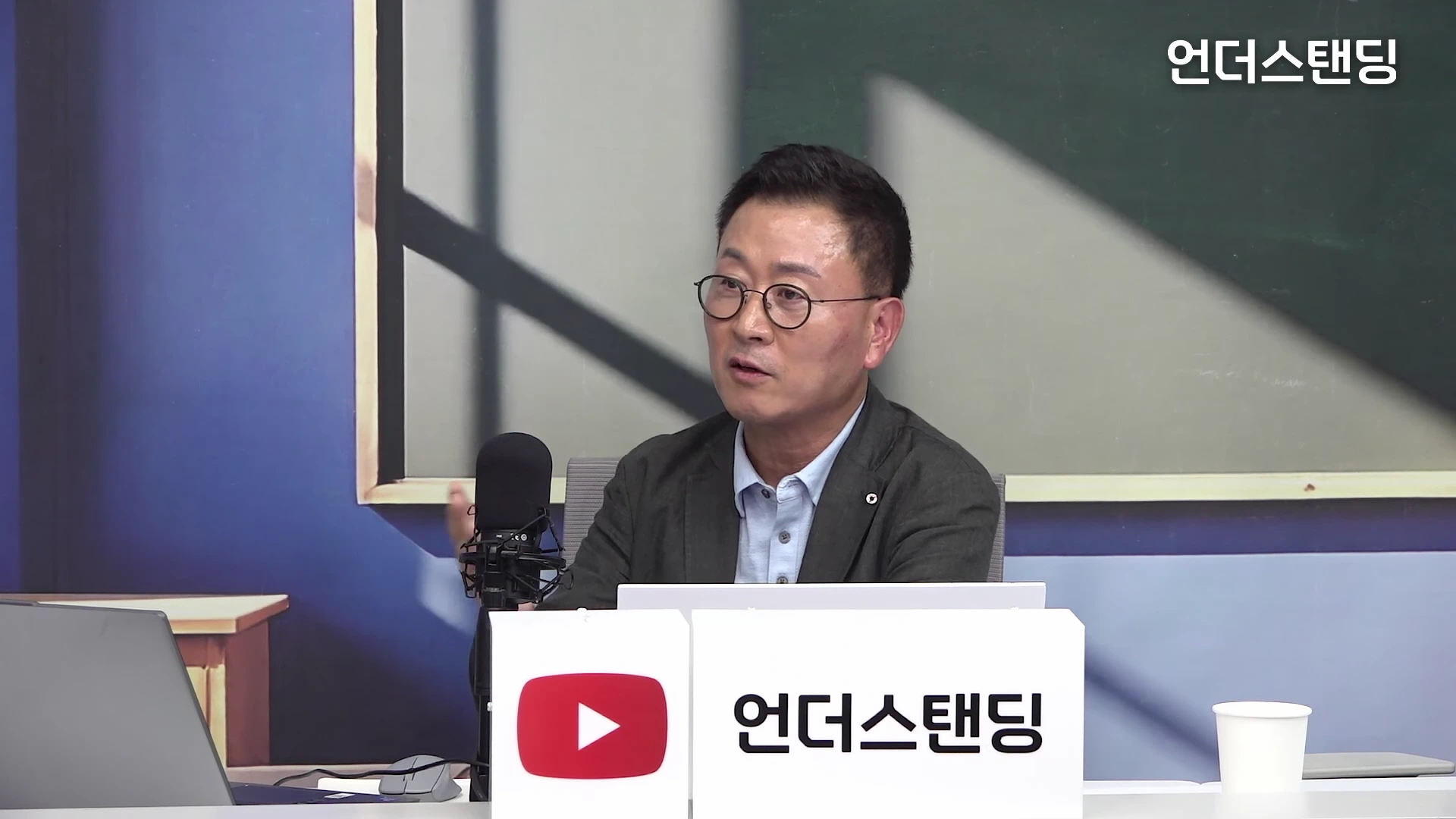 안경을 쓴 남성이 스튜디오 책상 앞에 앉아 마이크를 앞에 두고 이야기를 나누고 있는 모습