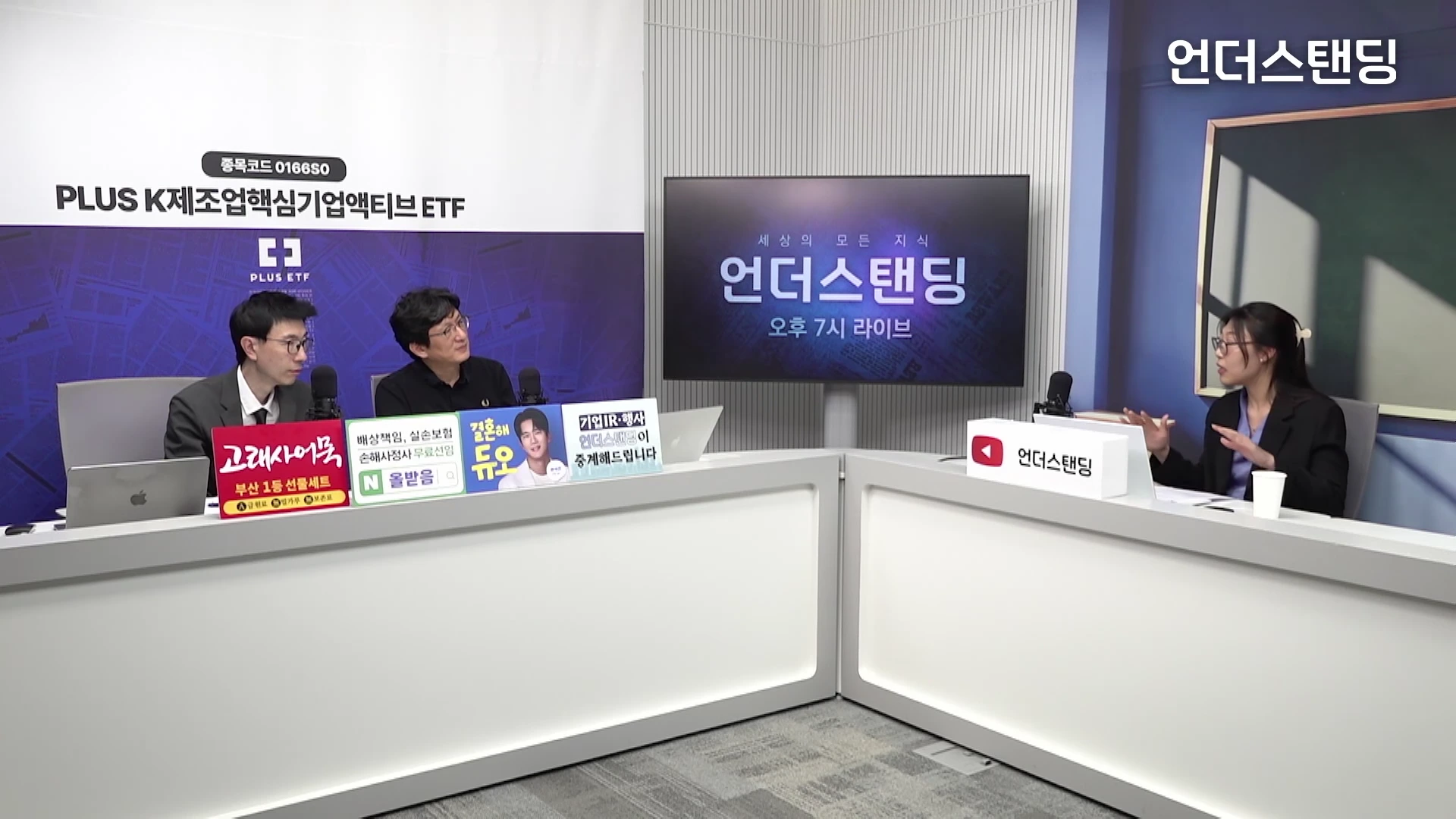 두 명의 남성과 한 명의 여성이 스튜디오 테이블에 앉아 대화를 나누고 있는 모습입니다. 각자의 앞에는 마이크와 다양한 홍보 문구가 적힌 작은 패널들이 놓여 있습니다.