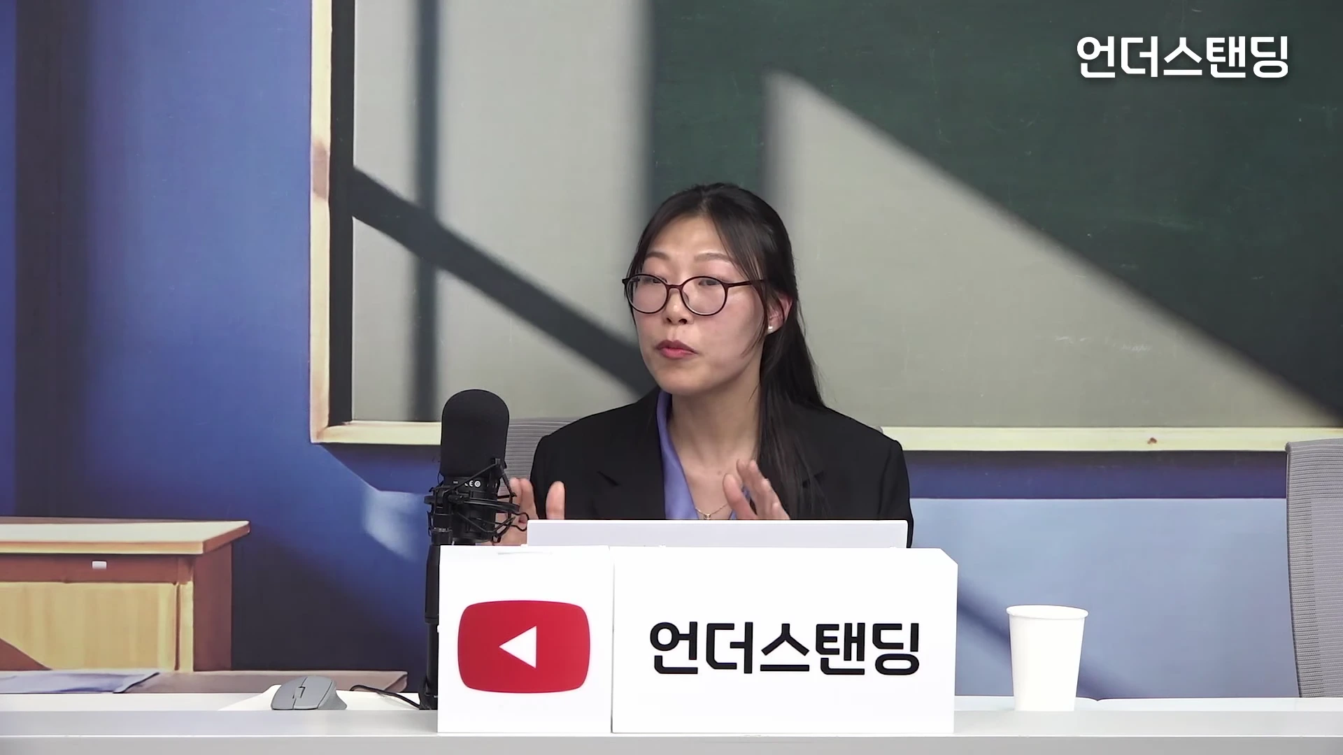 안경을 쓴 여성이 스튜디오에서 마이크 앞에 앉아 대화를 나누고 있다.