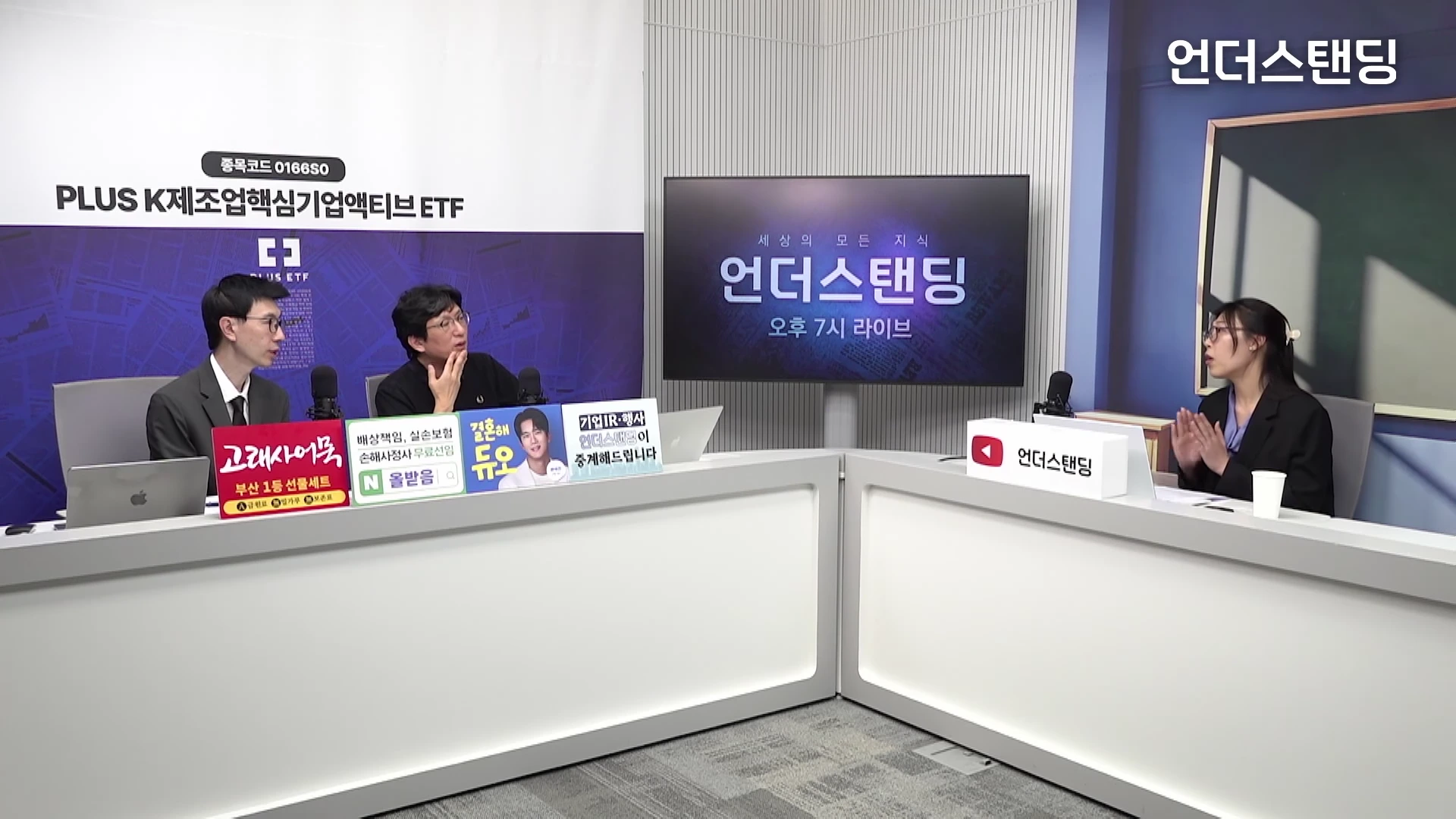 스튜디오 책상에 앉은 세 사람이 마이크를 앞에 두고 대화를 나누고 있는 모습으로, 중앙에는 대형 모니터가 설치되어 있고 책상 위에는 광고 문구가 적힌 작은 패널들이 놓여 있습니다.