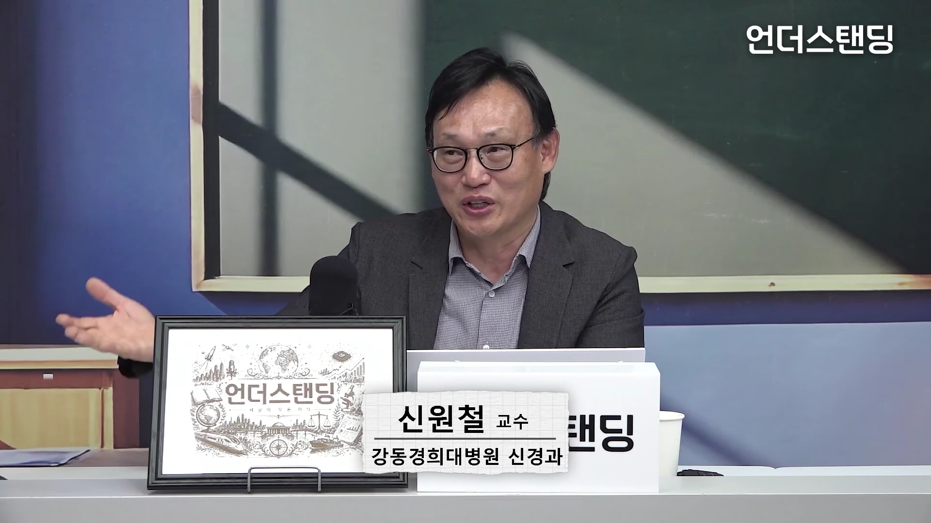 안경을 쓴 중년 남성이 스튜디오 테이블 앞에 앉아 손을 들어 설명하고 있는 모습으로, 테이블 앞에는 이름과 소속이 적힌 명패와 로고가 새겨진 액자가 놓여 있습니다.