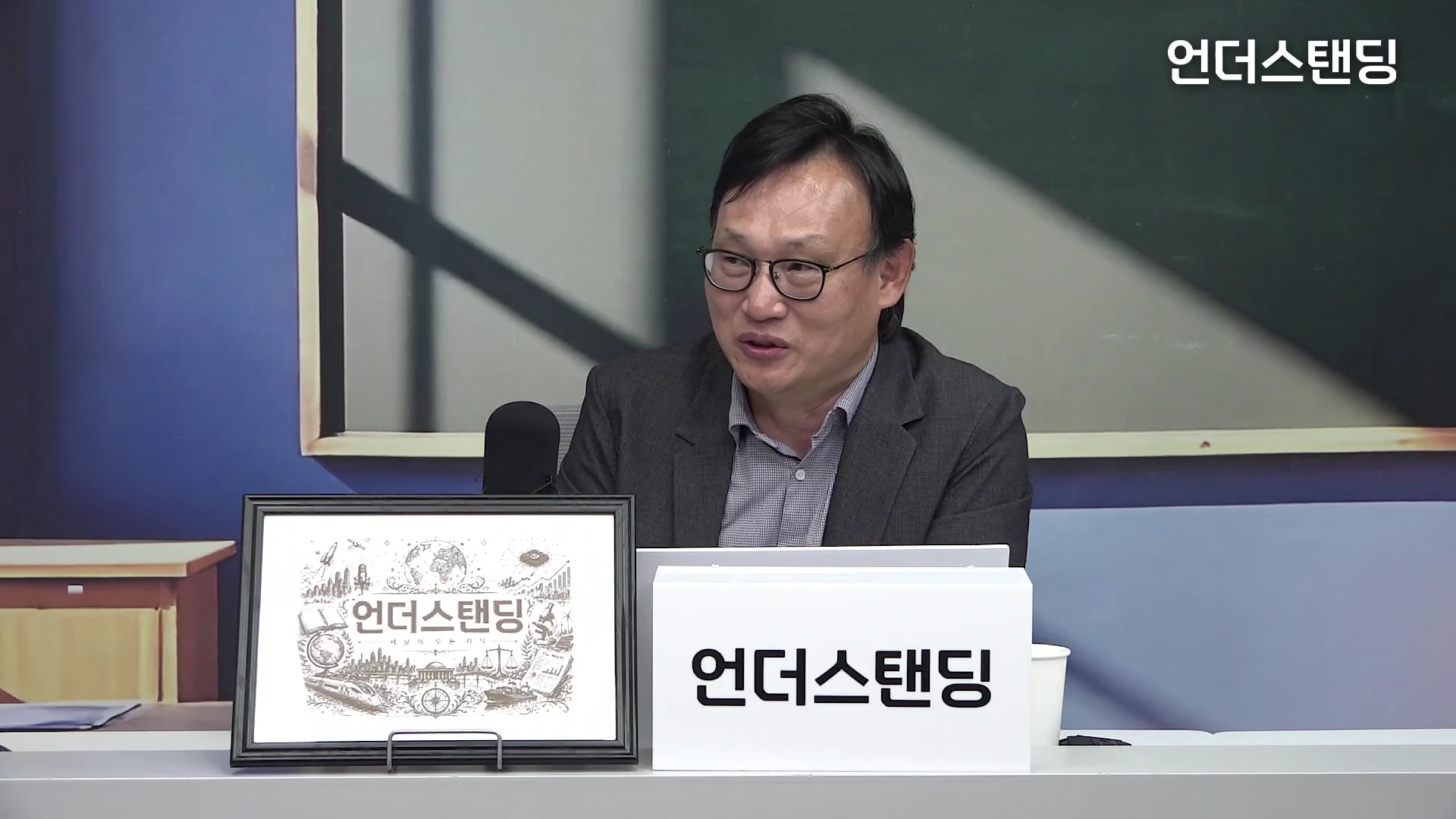 안경을 쓴 중년 남성이 스튜디오 책상에 앉아 마이크를 앞에 두고 인터뷰를 진행하고 있으며, 앞쪽에는 방송 로고가 들어간 액자와 이름표가 놓여 있다.