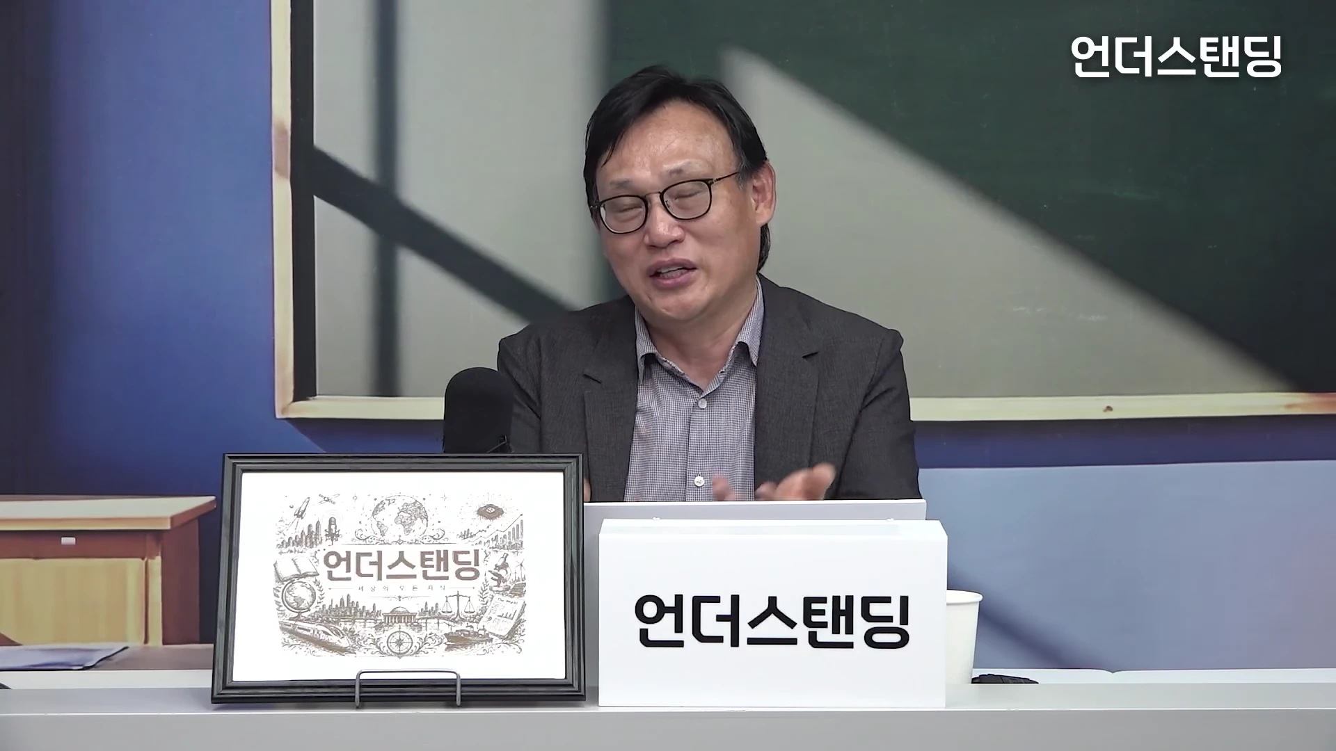 안경을 쓴 남성 출연자가 스튜디오 책상 앞에 앉아 이야기하고 있으며, 앞에는 로고가 새겨진 액자와 표지판이 놓여 있다.