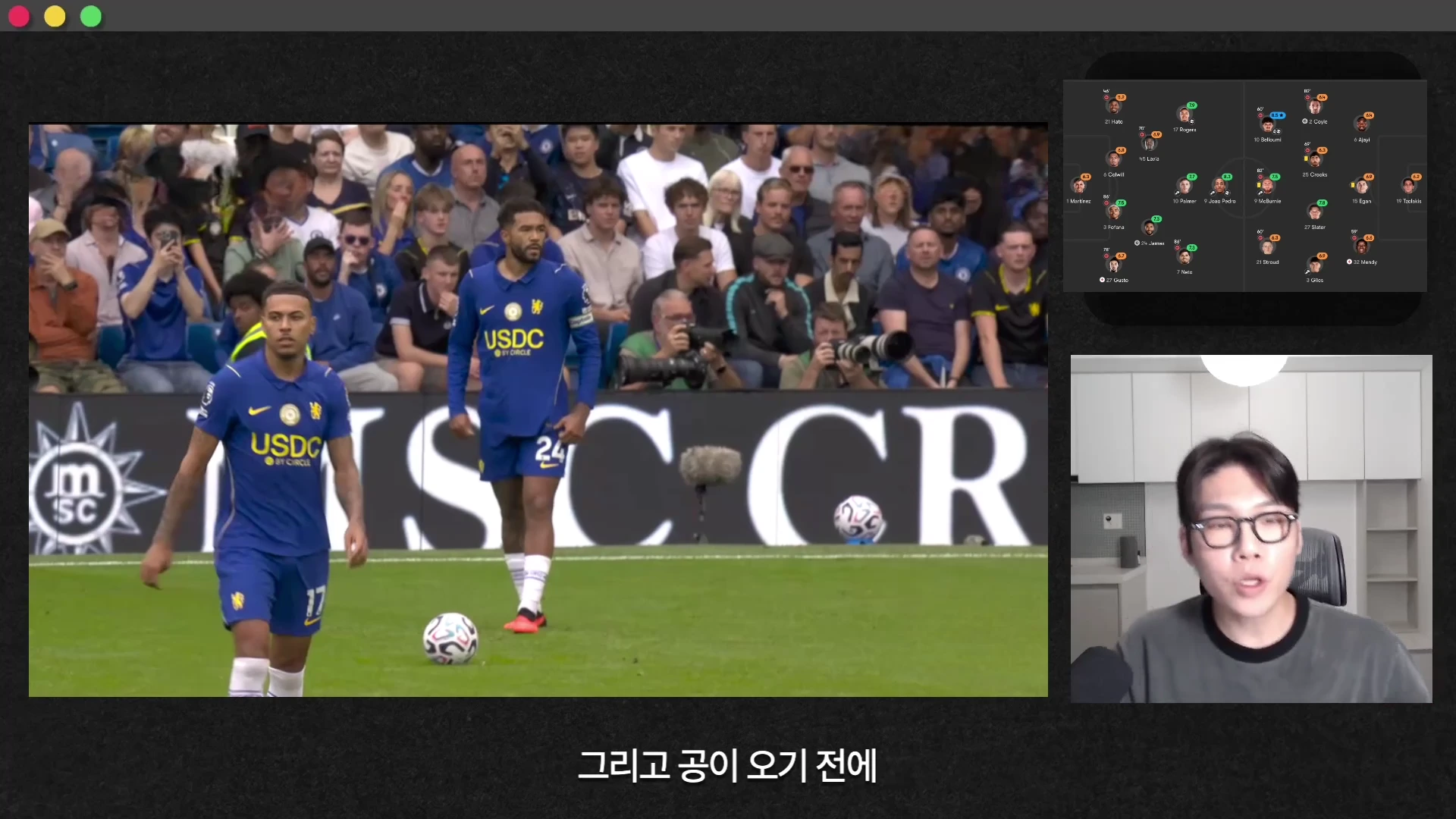 축구 경기장에서 파란색 유니폼을 입은 선수들이 서 있고, 화면 우측 상단에는 경기 정보 그래픽이, 우측 하단에는 안경을 쓴 남성이 출연하는 영상의 한 장면입니다.