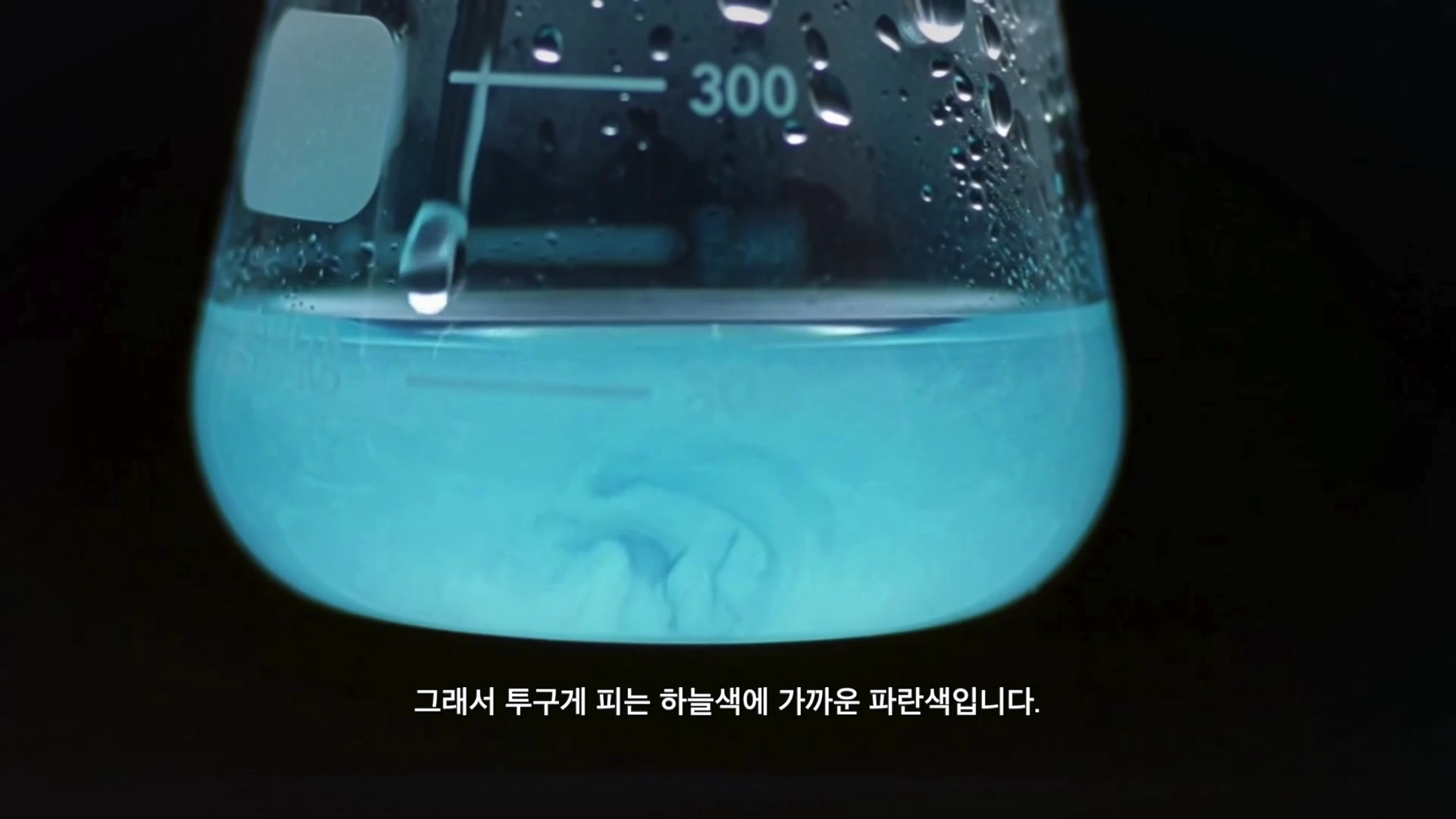 투명한 삼각 플라스크 안에 밝은 하늘색 액체가 담겨 있고, 액체 표면에 물방울이 떨어지는 모습이 클로즈업된 화면.
