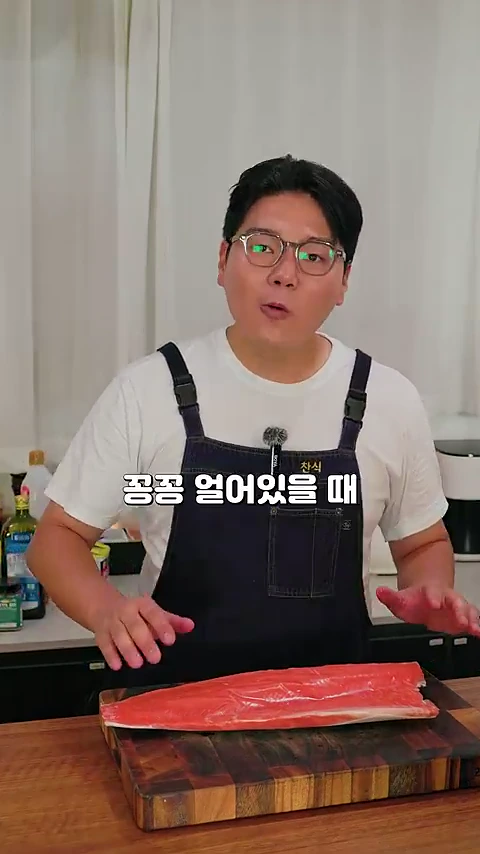 안경을 쓴 요리사가 흰색 티셔츠 위에 앞치마를 입고 도마 위에 놓인 냉동 연어 필렛 앞에 서서 설명하고 있는 모습.