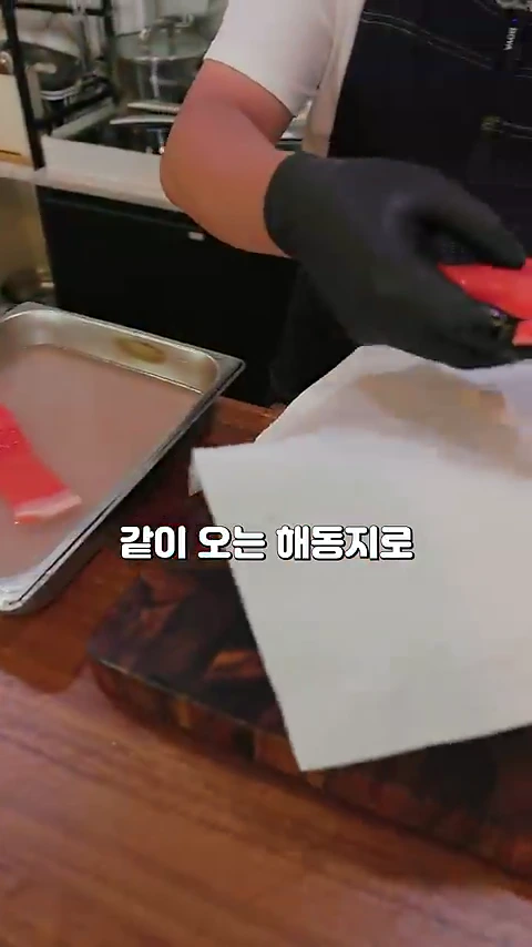 검은색 조리용 장갑을 낀 요리사가 나무 도마 위에서 해동지를 사용해 연어의 물기를 닦아내고 있는 모습입니다.