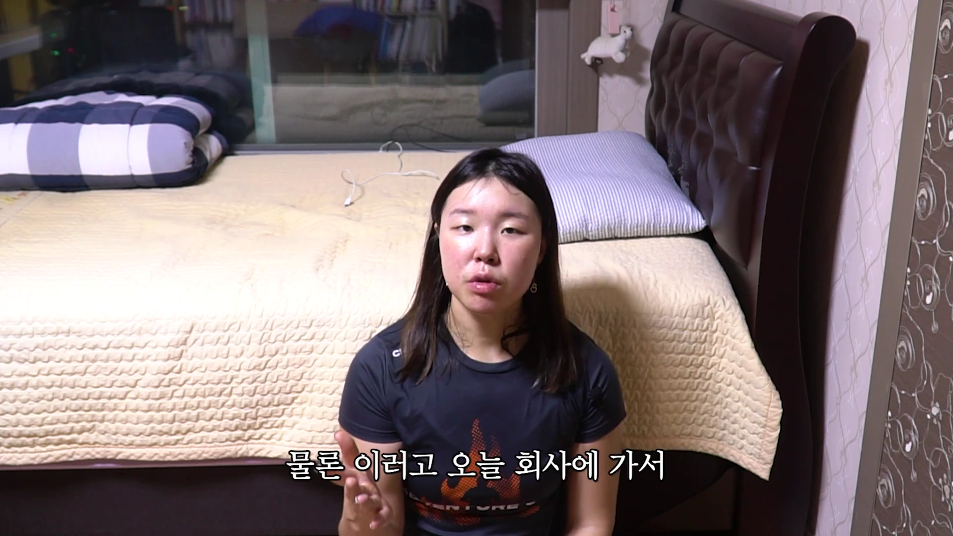 실내 침대 앞에 앉아 카메라를 보며 이야기를 나누고 있는 긴 머리의 여성.