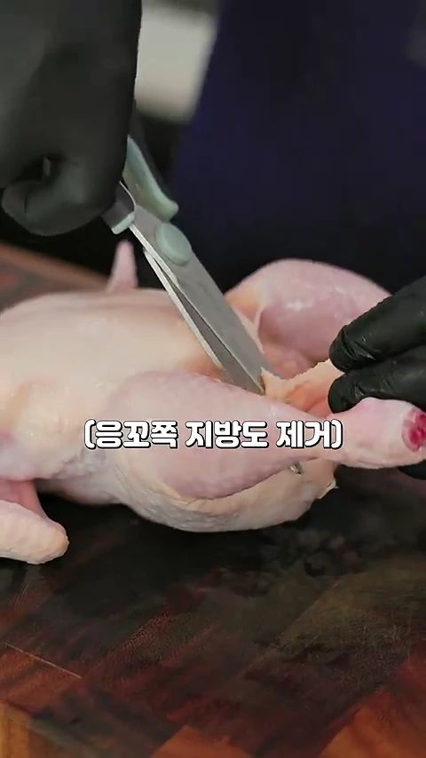검은색 장갑을 낀 손이 주방 가위로 생닭의 꼬리 부분 지방을 제거하는 모습
