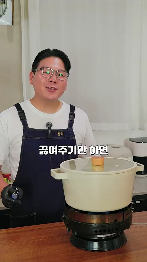 안경을 쓴 요리사가 흰색 티셔츠에 앞치마를 두르고 냄비가 놓인 가스레인지 앞에서 이야기하고 있다.