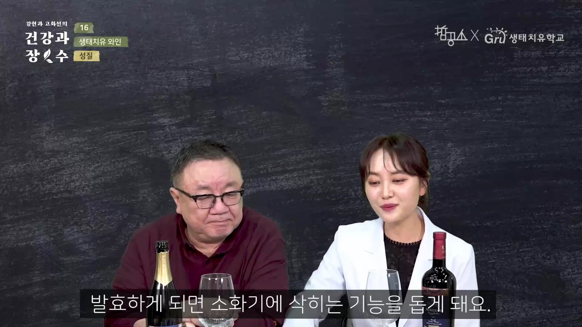 두 사람이 와인병과 와인잔이 놓인 테이블 앞에 앉아 이야기를 나누고 있는 방송 화면입니다.