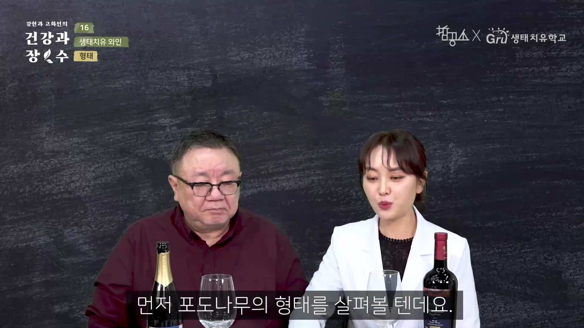 두 사람이 어두운 배경 앞에서 대화하고 있으며, 테이블 위에는 와인병과 와인 잔이 놓여 있다.