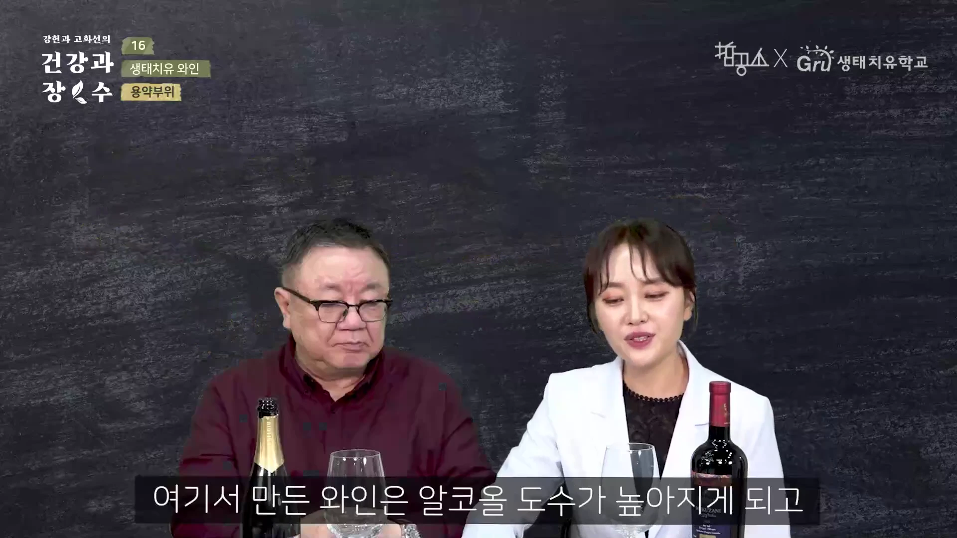 짙은 배경의 스튜디오에서 중년 남성과 젊은 여성이 와인병과 와인잔이 놓인 테이블 앞에 앉아 대화하고 있는 모습.