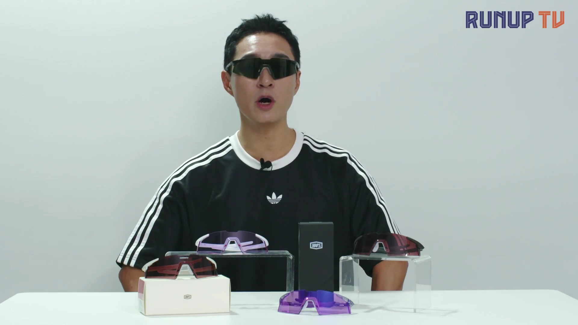 흰색 배경의 스튜디오에서 검은색 반팔 티셔츠를 입고 고글을 착용한 남성이 말을 하고 있으며, 앞쪽 테이블 위에는 다양한 색상의 고글과 전용 케이스가 진열되어 있습니다.