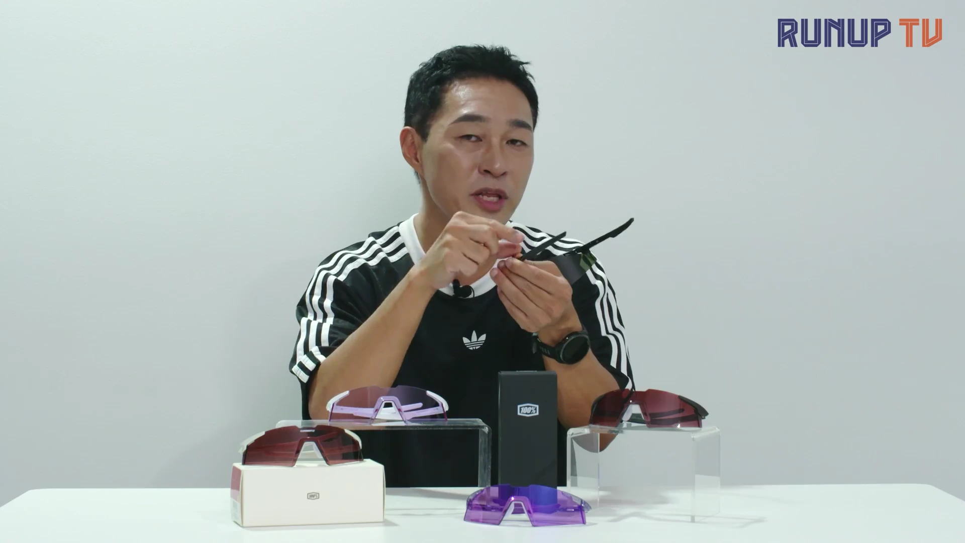 한 남성이 흰색 배경의 스튜디오에서 손에 든 검은색 고글의 힌지 부분을 보여주며 설명하고 있고, 앞쪽 테이블에는 다양한 색상의 고글 제품들이 놓여 있습니다.
