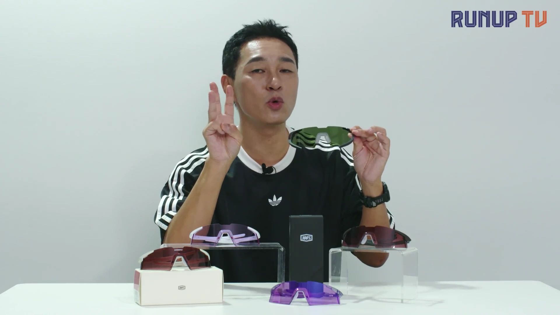 흰색 배경의 스튜디오에서 한 남성이 손가락으로 숫자 3을 표시하며 고글을 들고 설명하고 있고, 테이블 위에는 고글 세 개와 박스가 놓여 있습니다.
