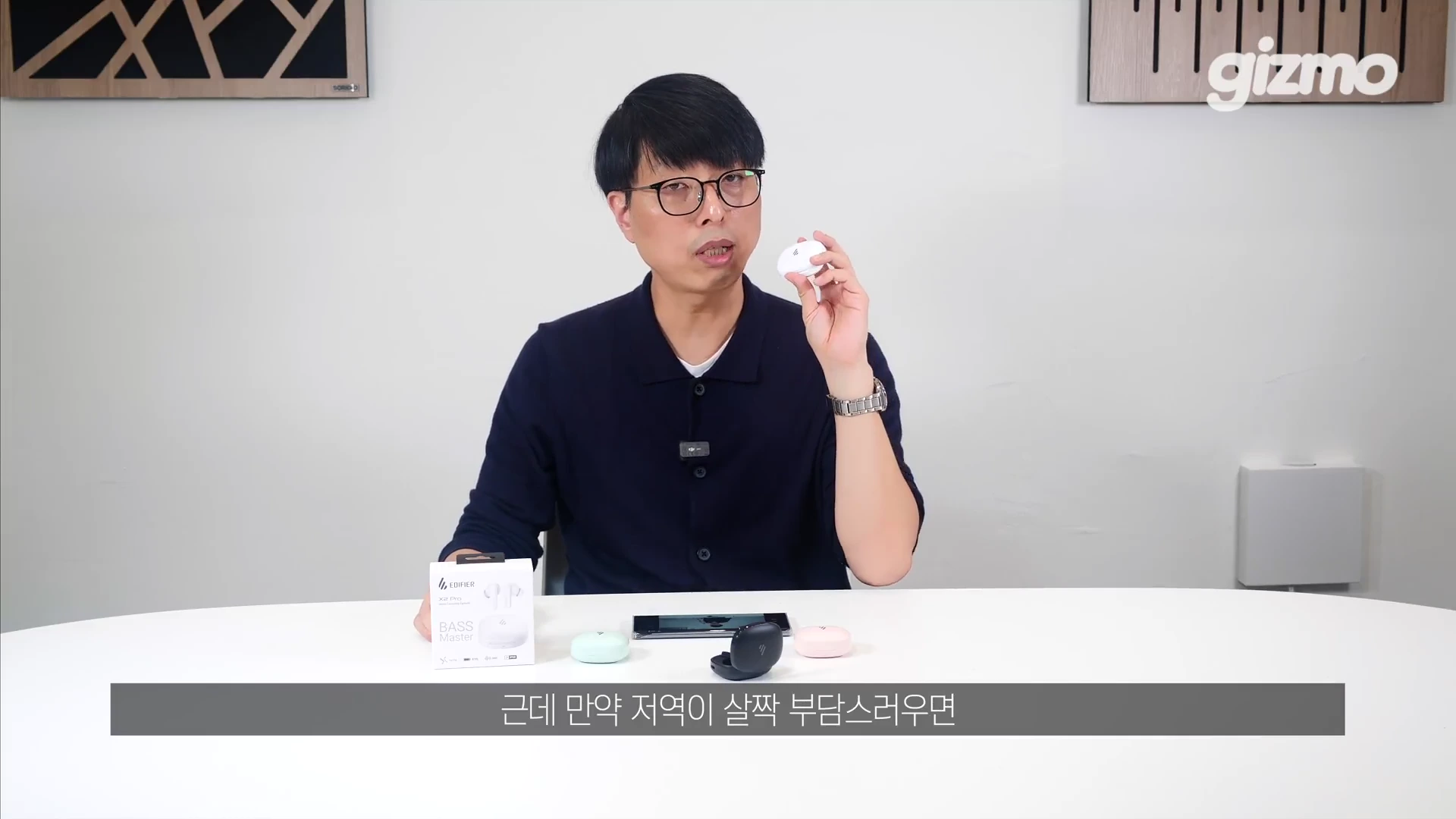 안경을 쓴 남성이 스튜디오 테이블에 앉아 무선 이어폰 충전 케이스를 손에 들고 설명하고 있으며, 테이블 위에는 제품 패키지와 다양한 색상의 이어폰 케이스들이 놓여 있습니다.