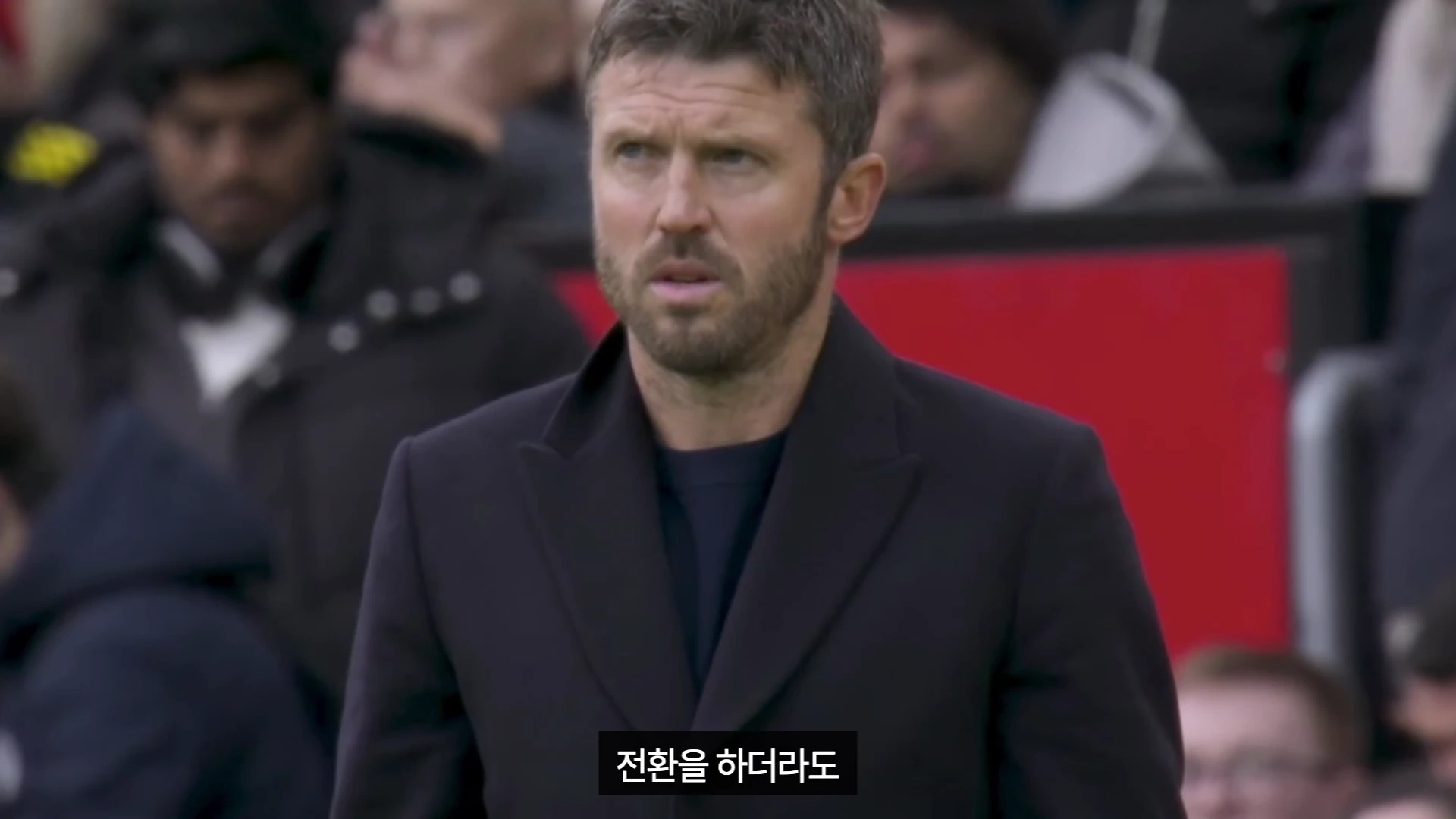 축구 경기장 터치라인에서 검은색 코트를 입고 근심 어린 표정으로 서 있는 마이클 캐릭 감독의 상반신 모습