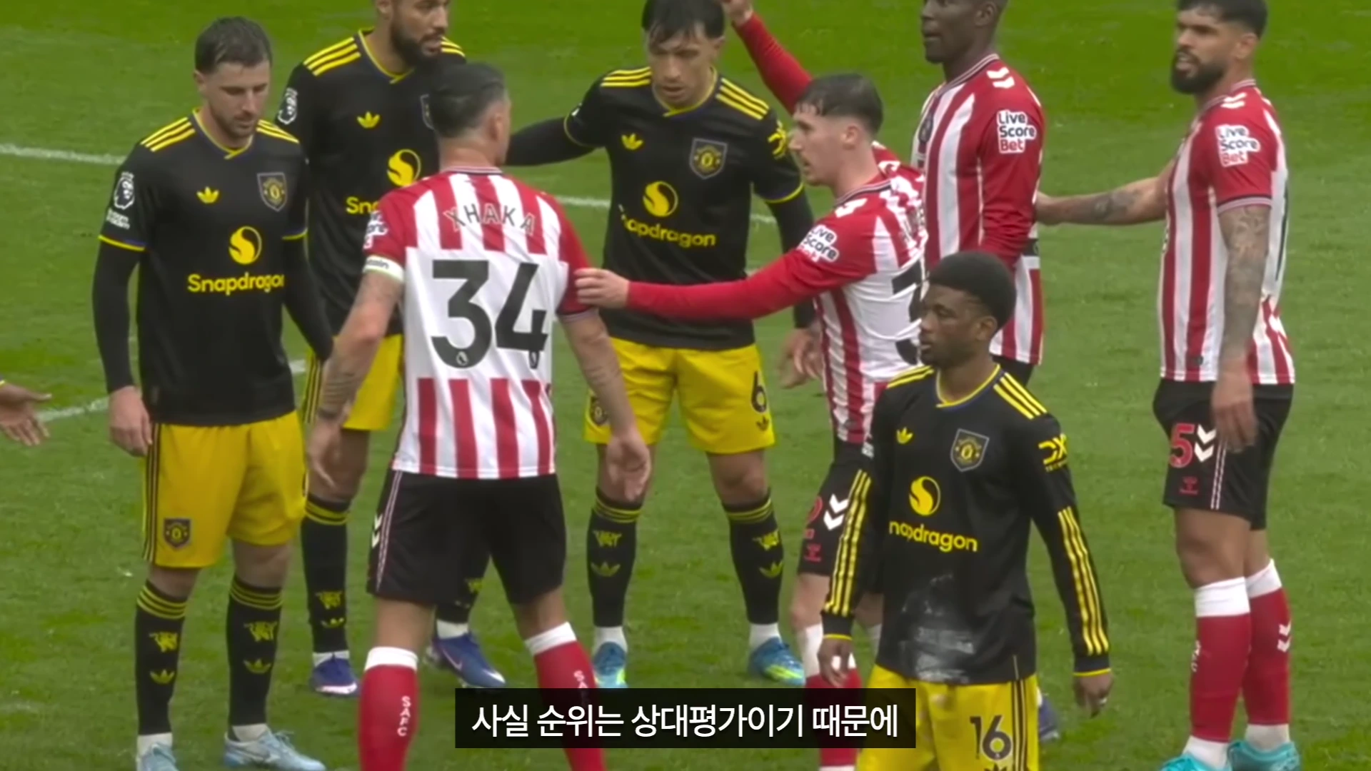 축구 경기장에서 검은색 원정 유니폼을 입은 선수들과 빨간색과 흰색 줄무늬 유니폼을 입은 선수들이 서로 엉켜 서 있는 모습.