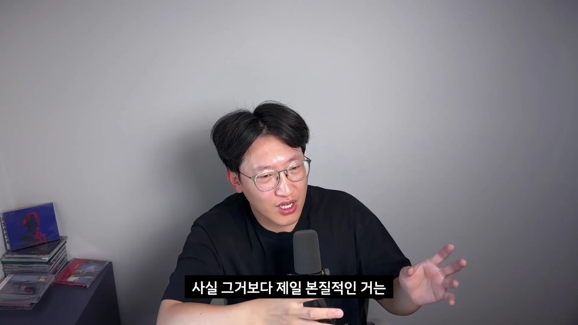 안경을 쓴 남성이 검은색 티셔츠를 입고 마이크 앞에 앉아 손짓하며 이야기하고 있으며, 화면 하단에는 '사실 그거보다 제일 본질적인 거는'이라는 한국어 자막이 표시되어 있습니다.