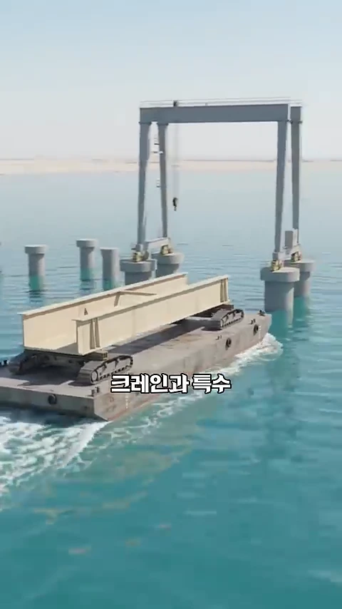 바다 위 교각들 사이로 대형 철제 구조물을 실은 바지선이 이동하고 있으며, 먼 배경에는 하늘과 맞닿은 수평선이 보인다.