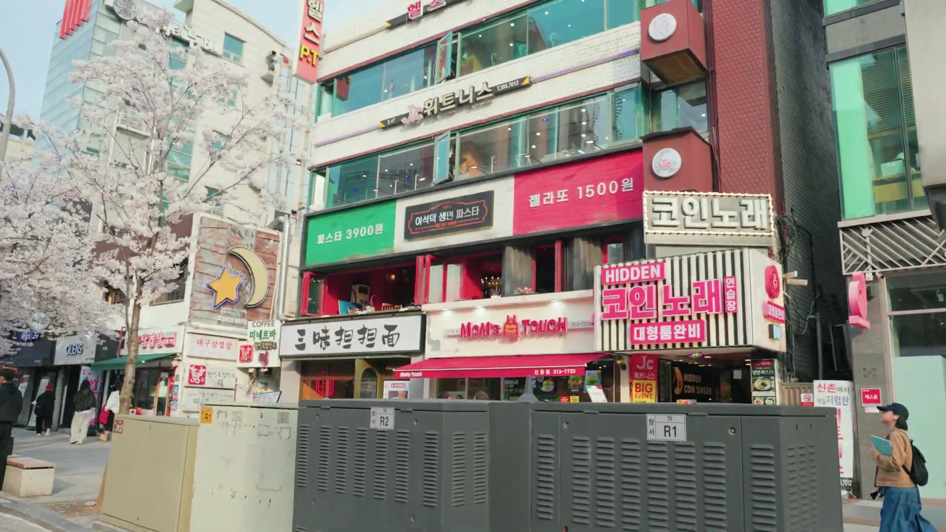 신촌 거리의 상가 건물 외관으로, 건물 전면에 '파스타 3900원'과 '젤라또 1500원'이라고 적힌 커다란 현수막이 붙어 있으며 주변에는 벚꽃이 피어 있다.