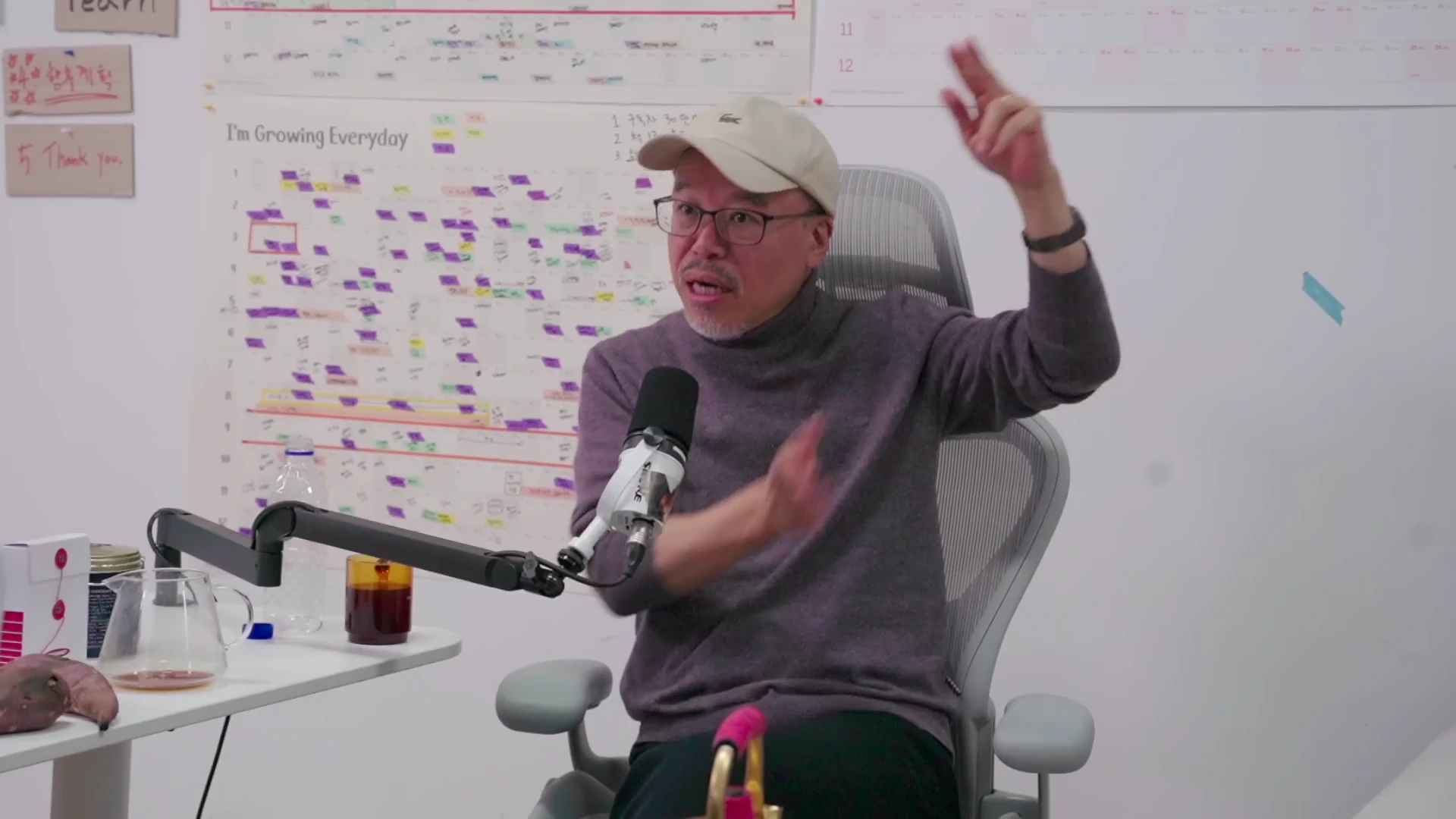 안경을 쓰고 야구 모자를 쓴 중년 남성이 스튜디오 마이크 앞에서 손을 들어 올리며 열정적으로 대화하고 있다. 배경에는 일정표가 붙은 화이트보드가 보인다.