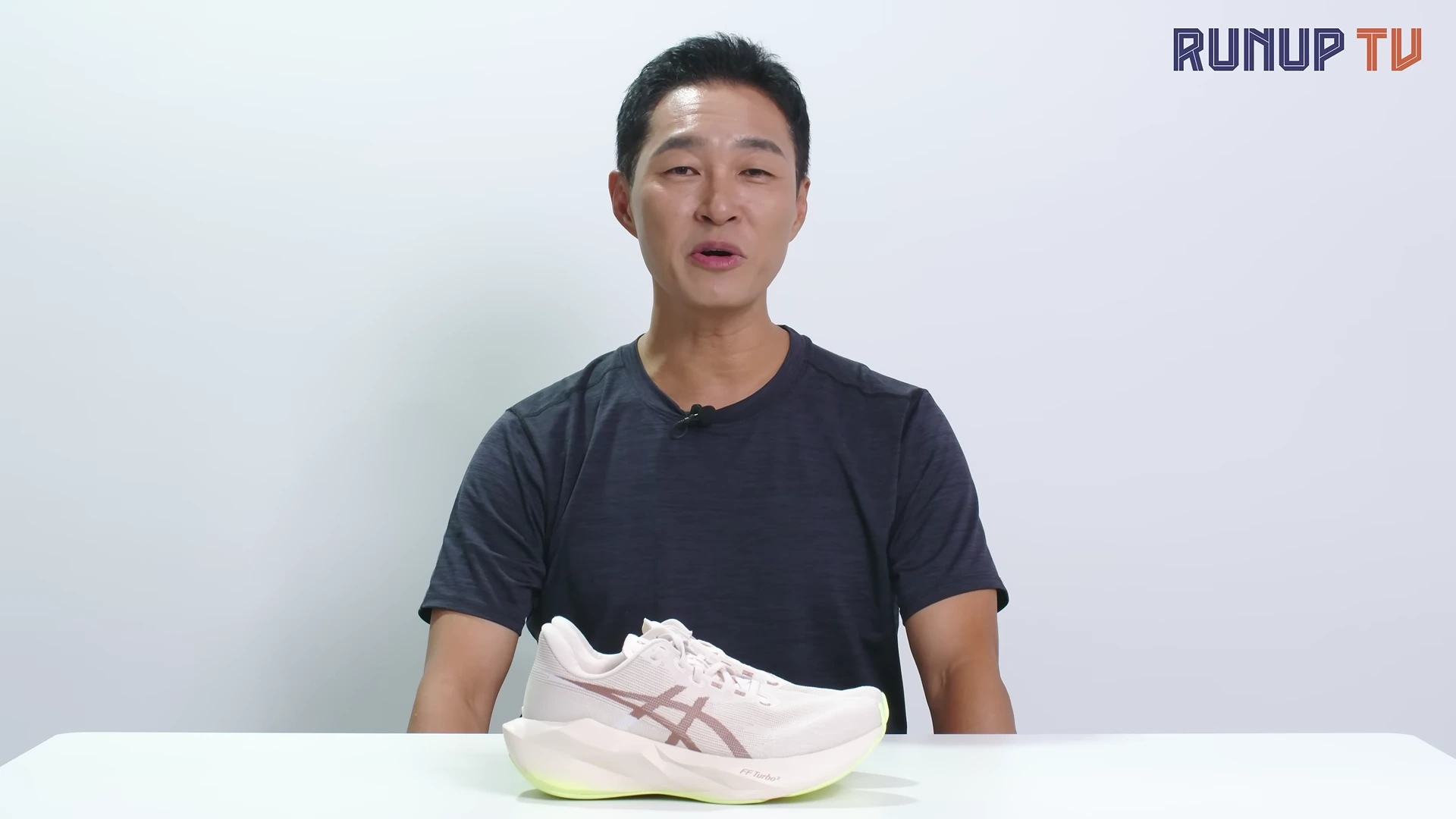 흰색 배경의 스튜디오에서 짙은 회색 티셔츠를 입은 남성이 앞에 놓인 밝은 베이지색 러닝화를 보며 이야기하고 있다.
