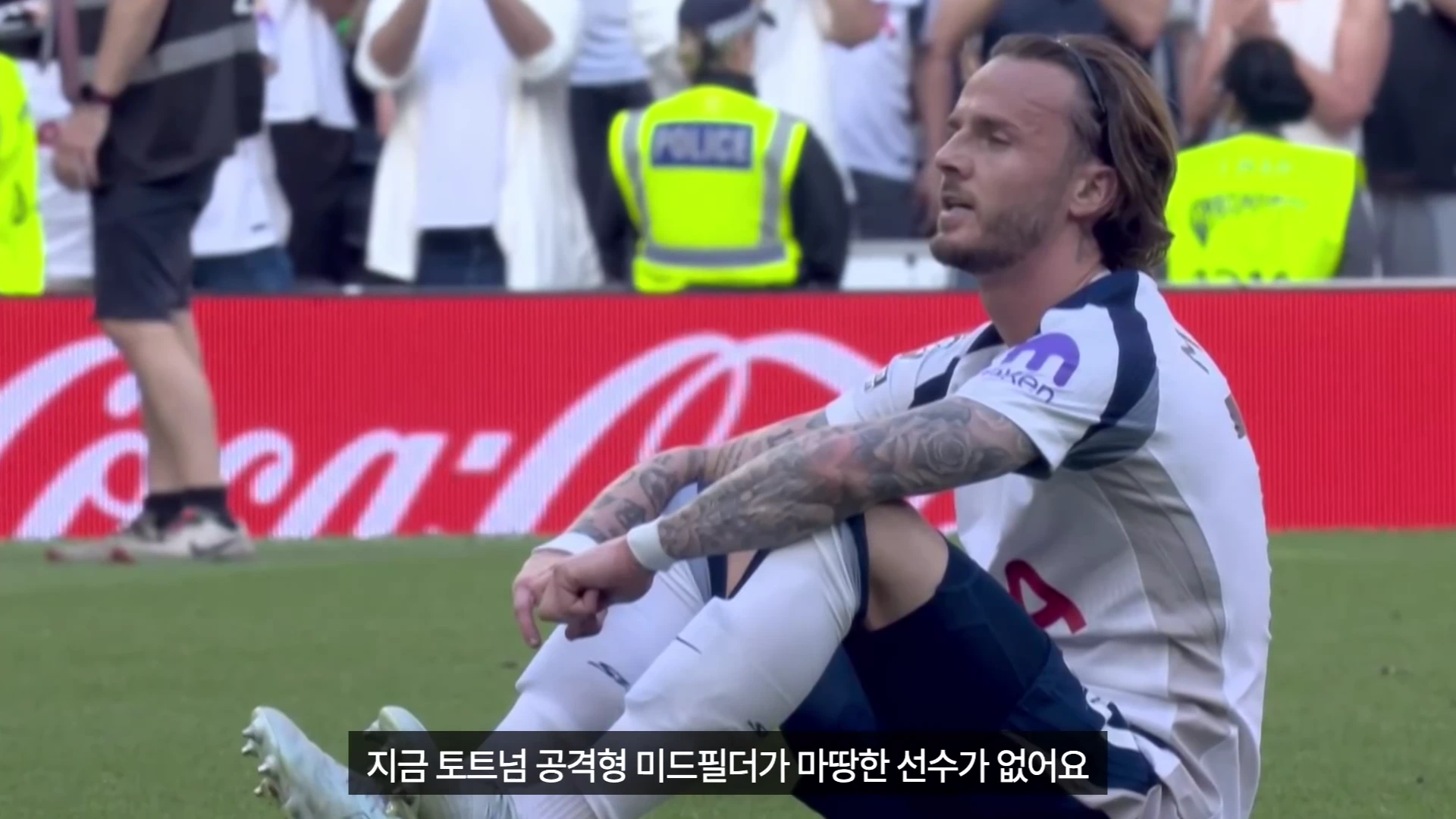 축구 경기장에서 흰색 토트넘 유니폼을 입은 한 선수가 잔디 위에 앉아 고개를 살짝 들고 있는 모습.