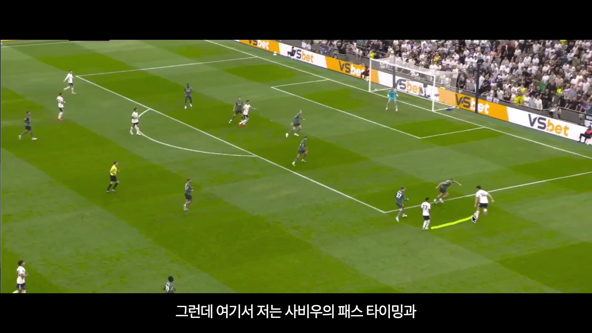 축구 경기 중 경기장 측면에서 한 선수가 다른 선수에게 패스를 보내는 장면을 담은 영상 캡처 화면으로, 노란색 화살표가 공의 이동 경로를 표시하고 있습니다.