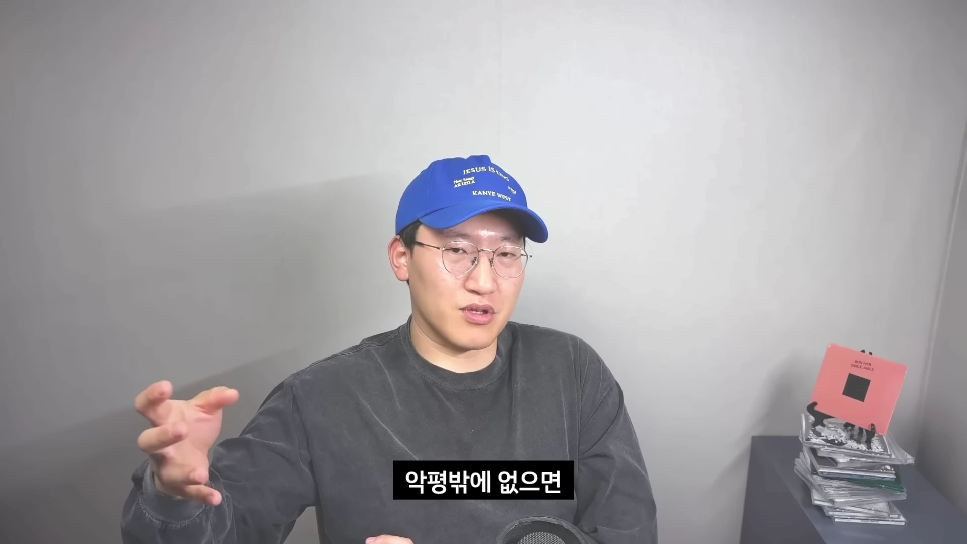 파란색 볼캡 모자와 안경을 쓴 남성이 회색 배경 앞에서 손짓하며 말을 하고 있는 모습