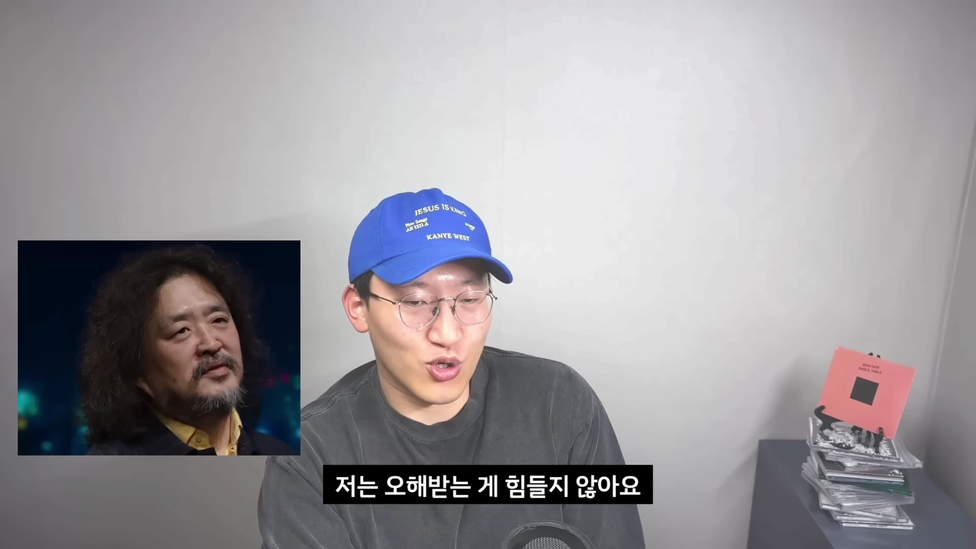 파란색 모자를 쓴 남성이 마이크 앞에서 이야기하고 있으며, 화면 왼쪽에는 긴 머리의 중년 남성이 나온 영상 장면이 삽입되어 있습니다.