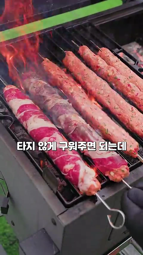 숯불 그릴 위에 긴 꼬치에 꽂힌 고기들이 나란히 놓여 있고, 한쪽에서 불꽃이 올라오며 고기가 구워지고 있는 모습.