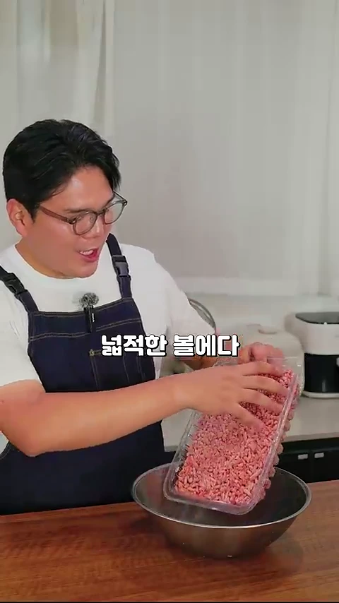 안경을 쓴 남성이 앞치마를 두르고 주방에서 투명한 플라스틱 용기에 담긴 다진 양고기를 커다란 스테인리스 볼에 붓고 있는 모습