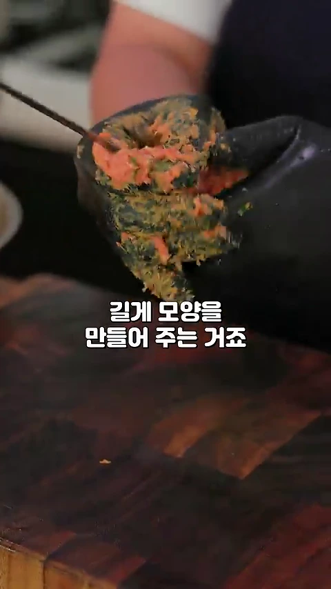 검은색 위생 장갑을 낀 손으로 양념된 고기 반죽을 꼬치에 감싸며 길게 빚고 있는 모습입니다.