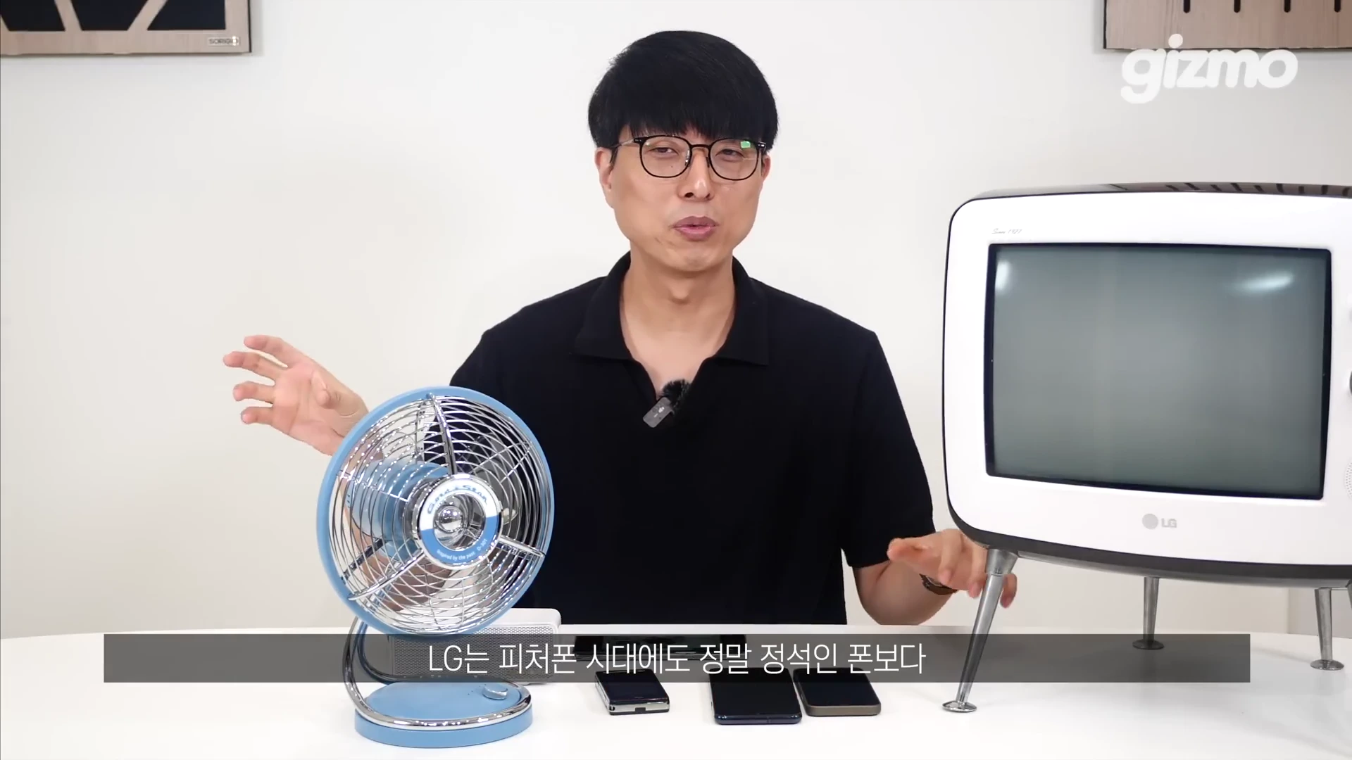 안경을 쓴 남성이 스튜디오 테이블 앞에 앉아 손짓하며 설명하고 있고, 테이블 위에는 금색 금성사 로고가 있는 레트로한 하늘색 선풍기와 레트로풍 TV, 몇 대의 스마트폰이 놓여 있습니다.
