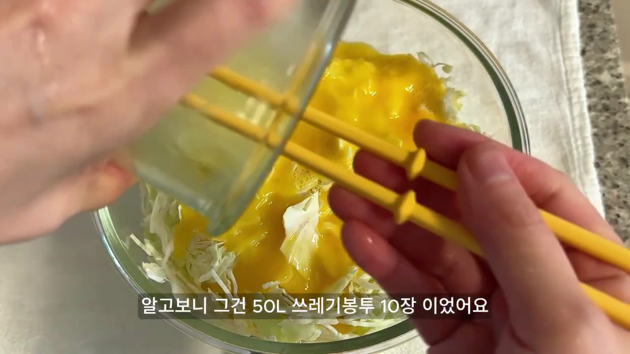 투명한 유리 그릇에 담긴 채 썬 양배추 위로 노란 계란물을 붓고 있는 손과 젓가락이 화면 하단에 텍스트와 함께 보인다.