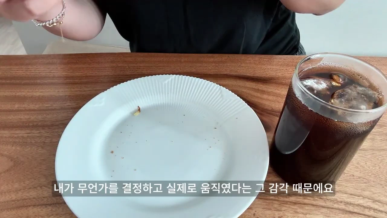 나무 테이블 위에 놓인 빈 접시와 얼음이 든 아이스 아메리카노 유리잔, 그리고 자막이 포함된 영상 화면.