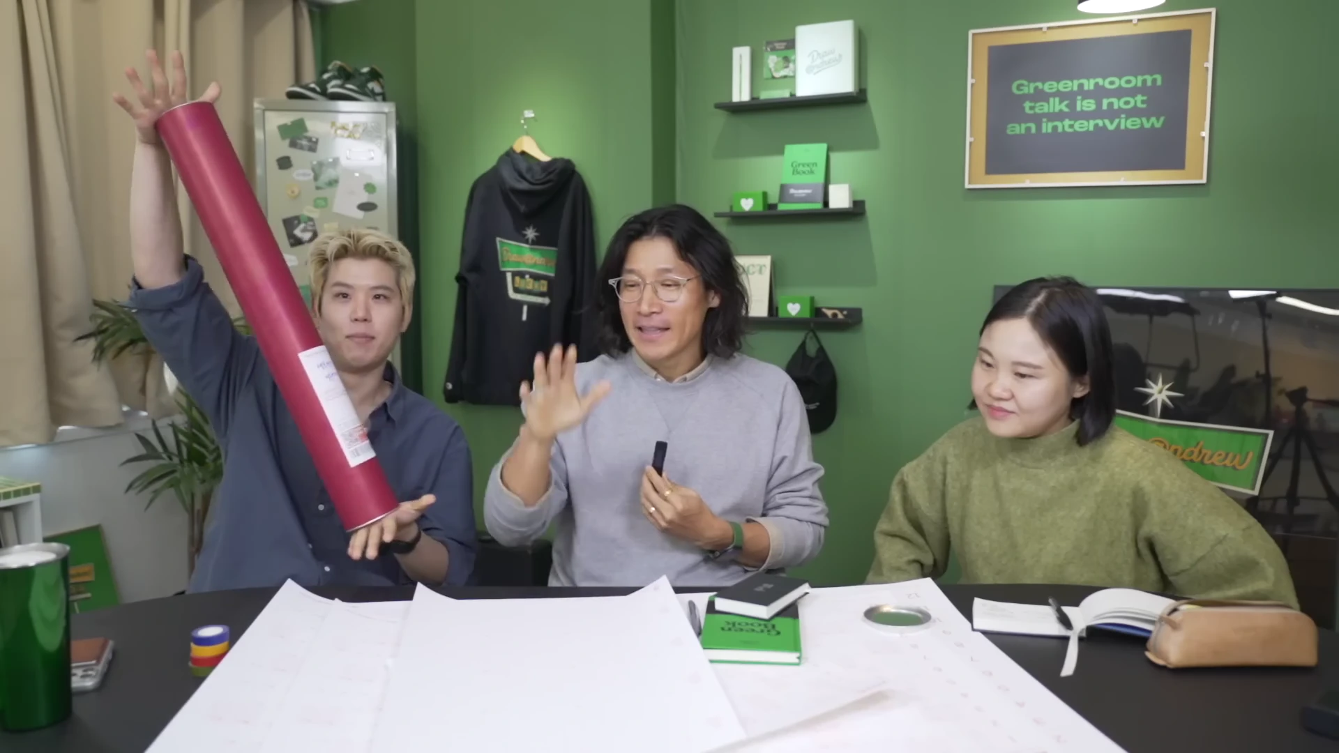 녹색 벽을 배경으로 한 스튜디오에서 세 사람이 테이블에 둘러앉아 대화하고 있으며, 한 남성이 긴 원통형 케이스를 높이 들어 보이고 있다.