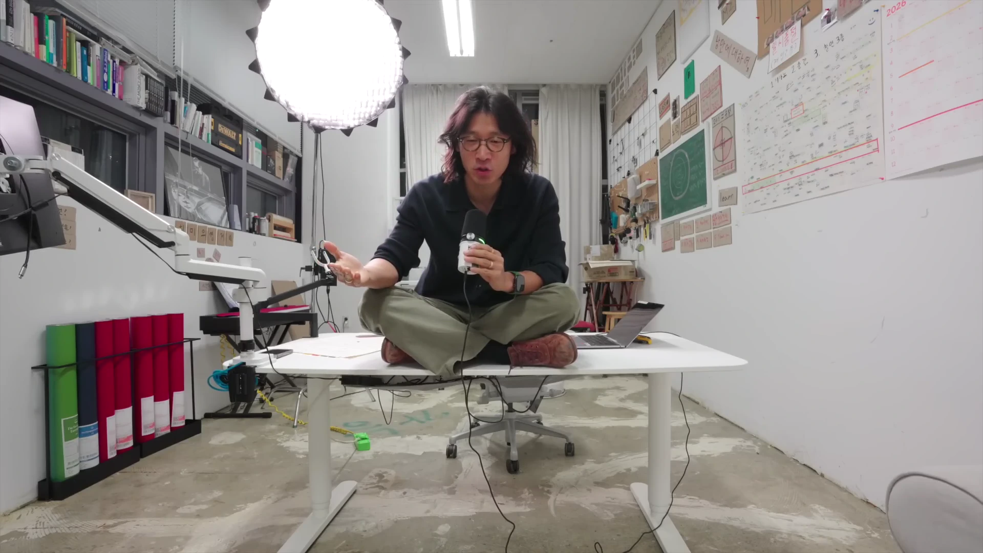 긴 머리의 남성이 작업실 책상 위에 책상다리를 하고 앉아 마이크를 든 채 이야기를 하고 있으며 배경에는 벽면 가득 기록물과 메모가 붙어 있다.